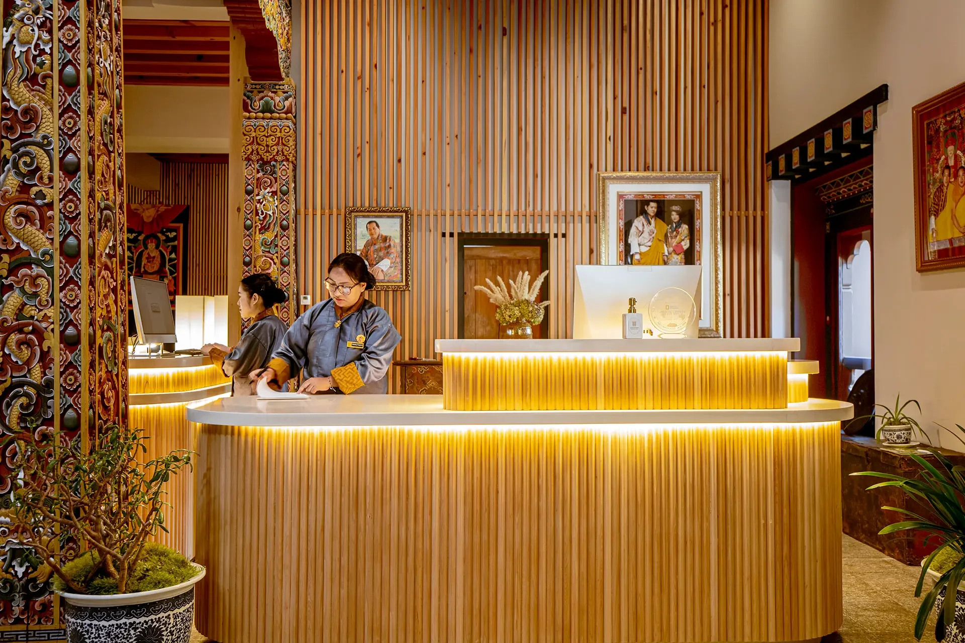 Hotel lobby reception desk with two staff members behind it, decorative wood paneling, framed pictures, indoor plants, and warm lighting.