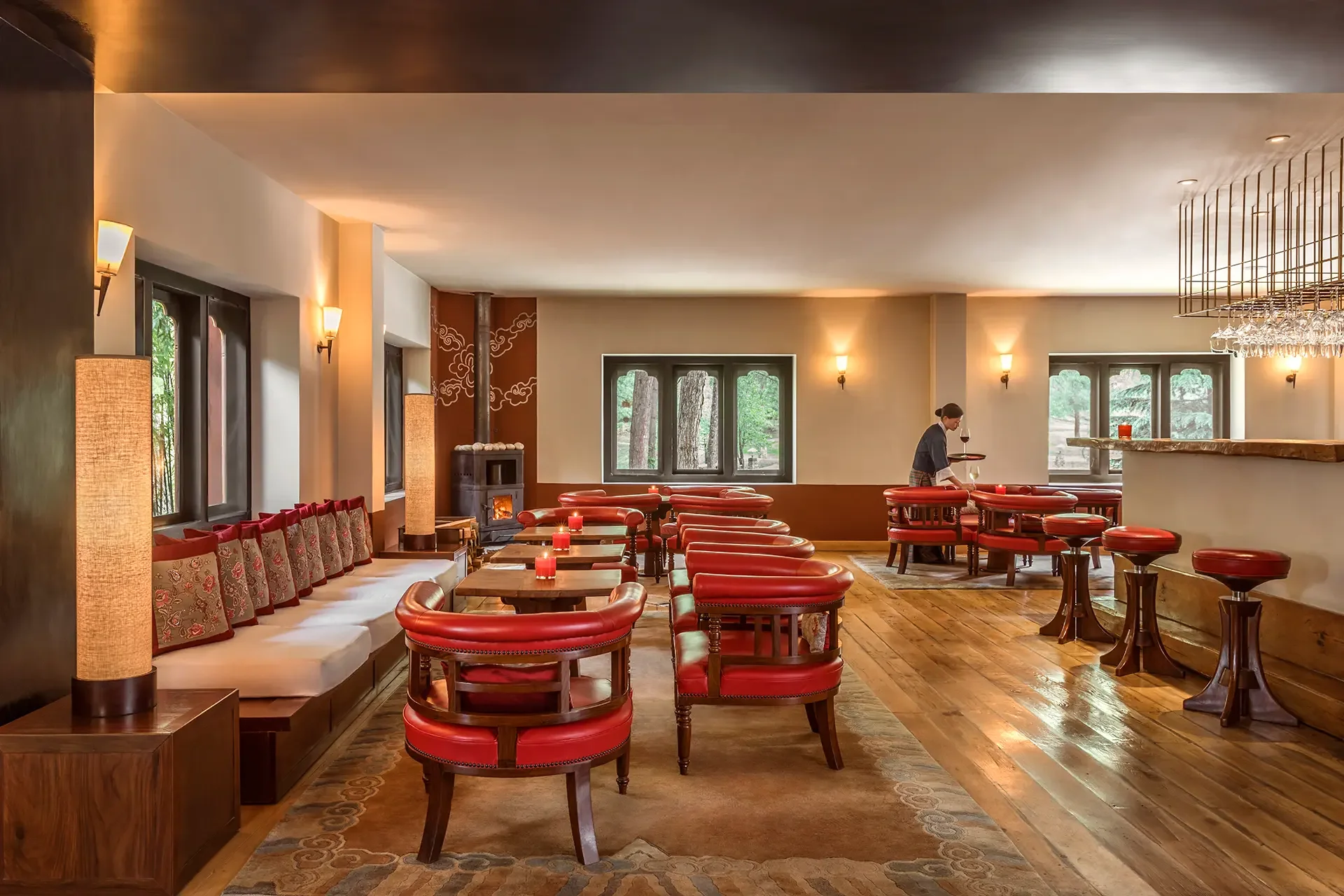 Interior of a cozy restaurant or lounge with wooden floors, red cushioned chairs, a long white cushioned bench with patterned pillows, tall windows showing trees outside, a wood-burning stove, and warm lighting from wall sconces and candles on tables. A woman is serving wine.