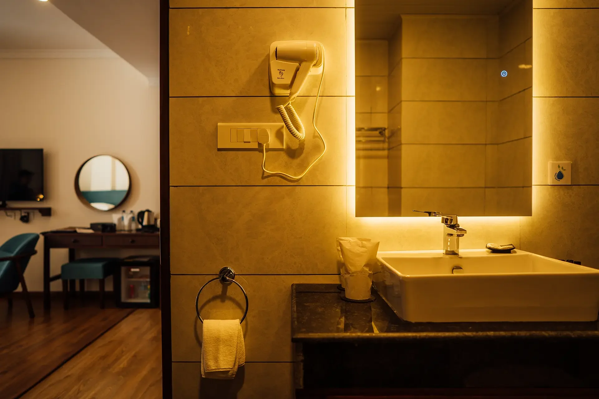 Bathroom with a sink, mirror, hair dryer, tissue holder, and a towel on a ring. The bathroom has a warm yellow lighting and a modern design.