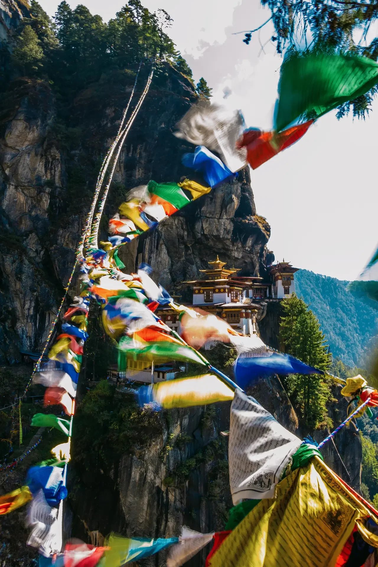 Colorful prayer flags hanging in front of a mountain landscape with a temple built into the rock face, surrounded by trees and blue sky.