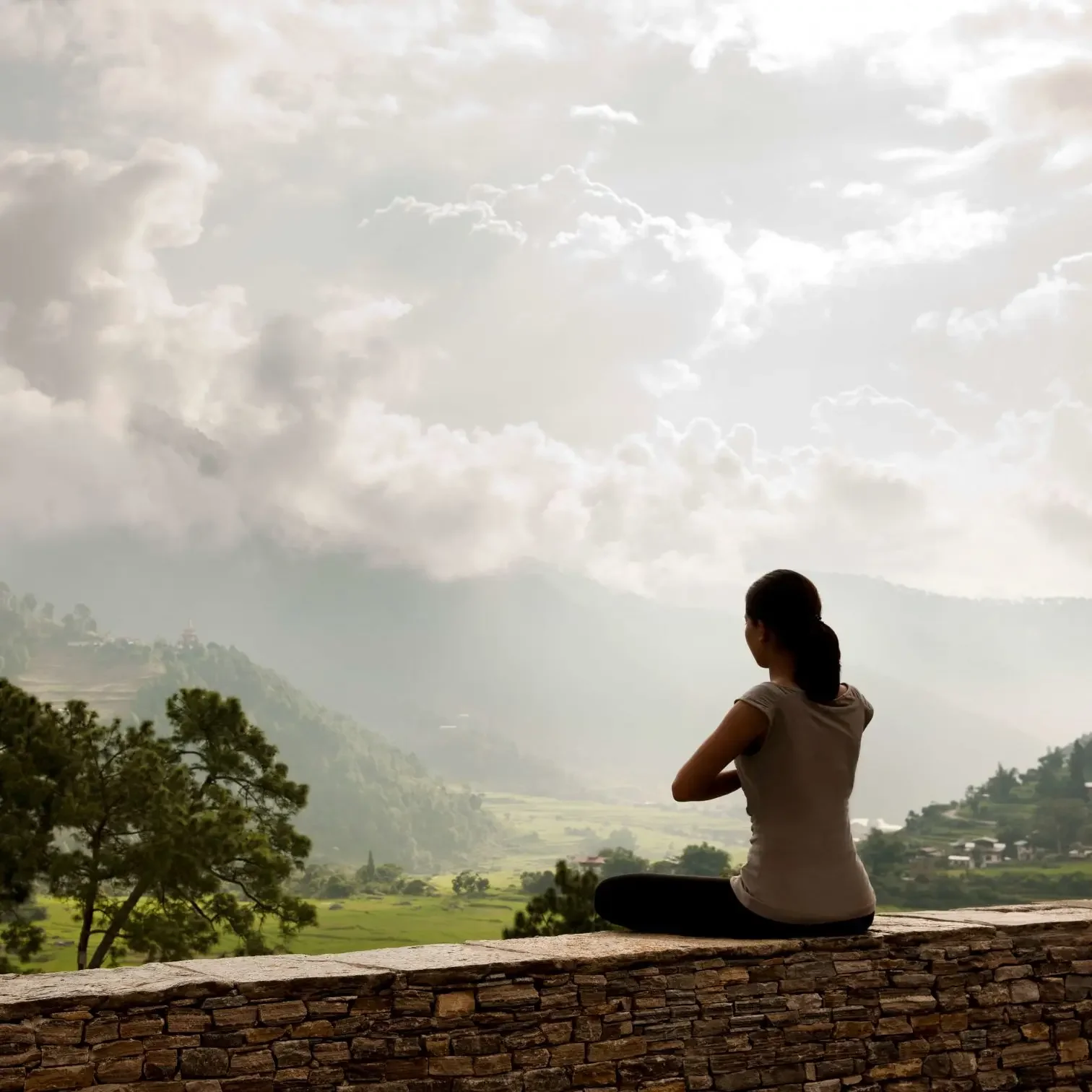 A woman sitting cross-legged on a stone ledge, looking at a scenic view of green hills, trees, and a cloudy sky during daytime.