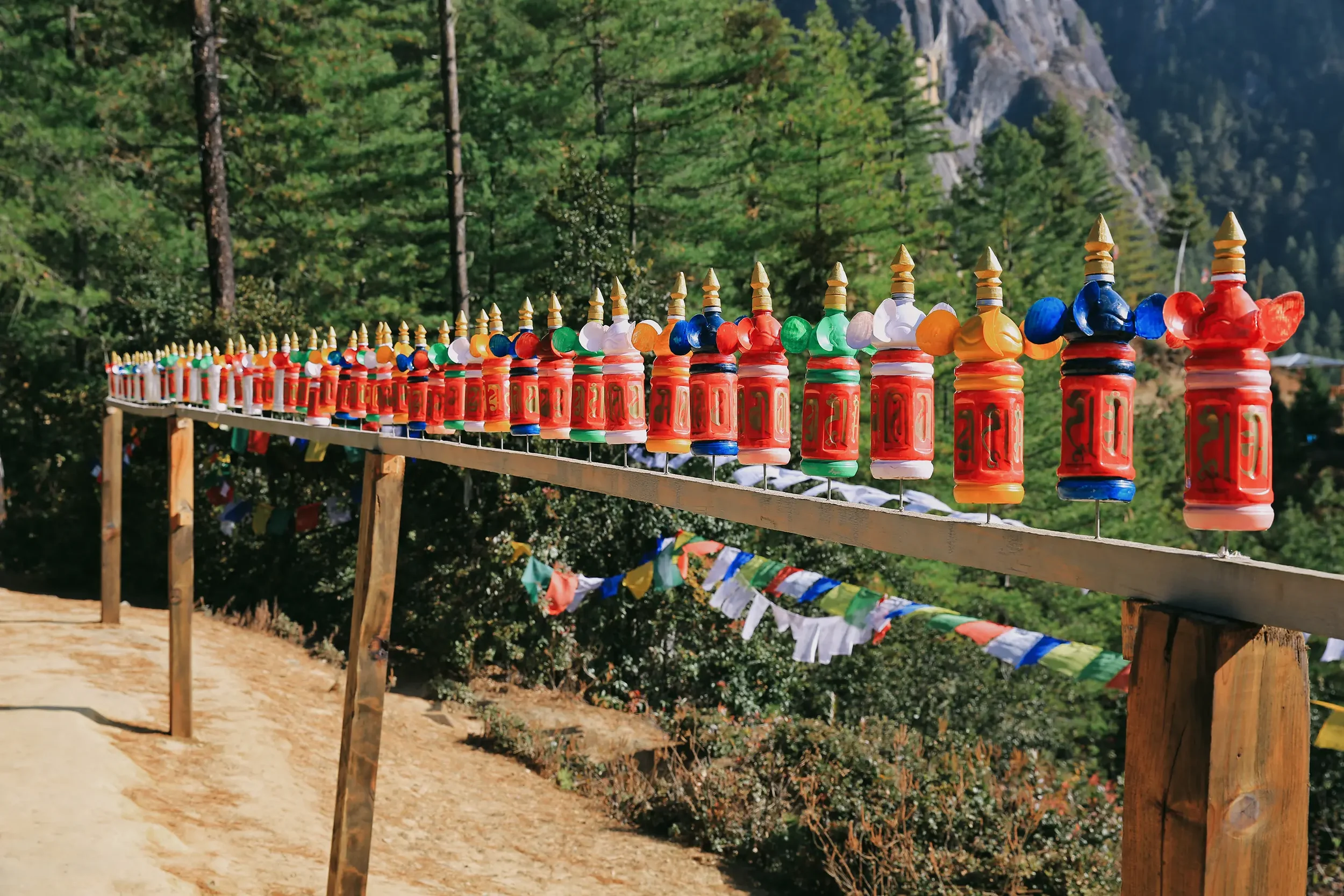 Colorful prayer flags and prayer wheels strung along a wooden fence in a mountainous forest setting.