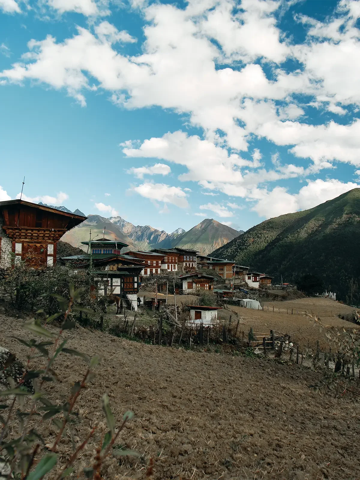 A mountain village with traditional wooden houses surrounded by lush green hills and mountains under a partly cloudy blue sky.