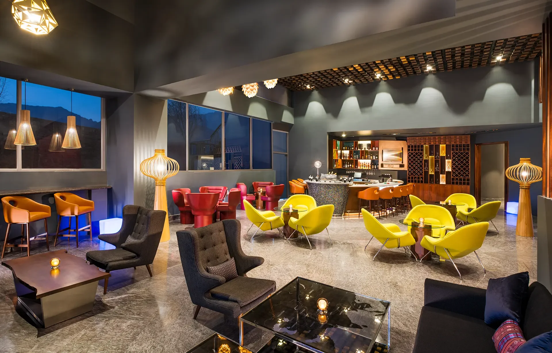 Interior of a modern bar and lounge area with vibrant yellow, red, and gray seating, decorated with wooden lighting fixtures, large windows, and a well-stocked bar in the background.