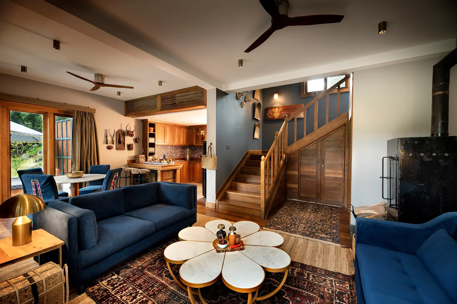 Living room with blue sofas, a white eclectic coffee table, a wooden staircase, a wood-paneled kitchen area, ceiling fans, and decorative wall art.
