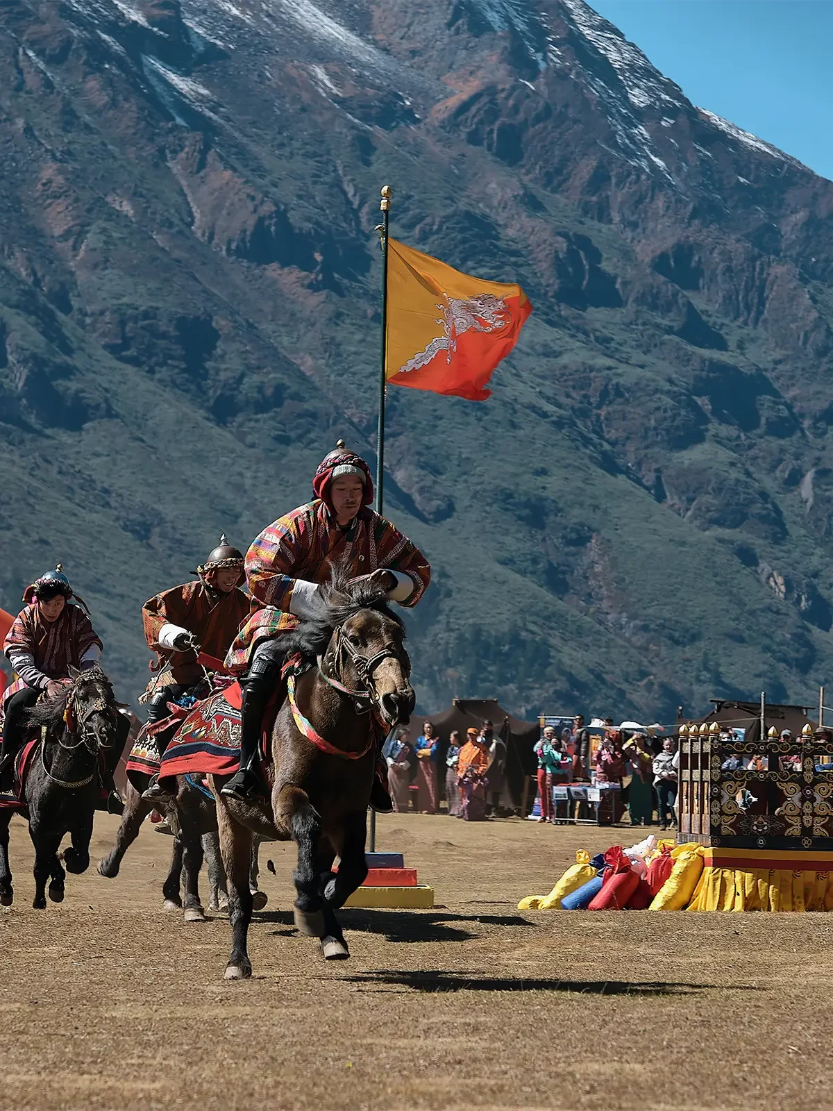 People are riding horses in a traditional race or event outdoors, with a mountainous landscape in the background, and a crowd of spectators and colorful decorations at the side.