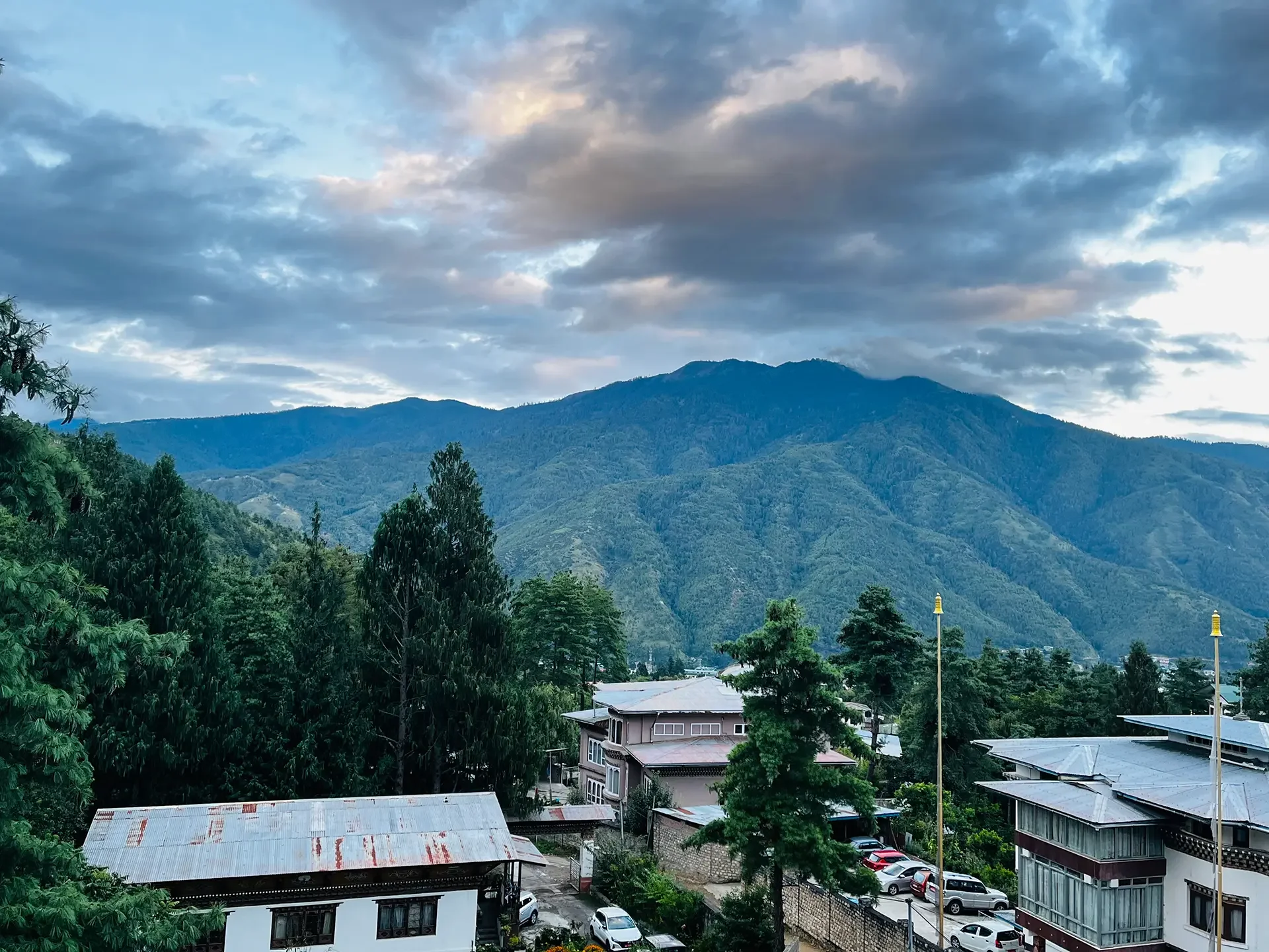 Mountain landscape with cloudy sky, lush green trees, and houses with metal roofs in the foreground.