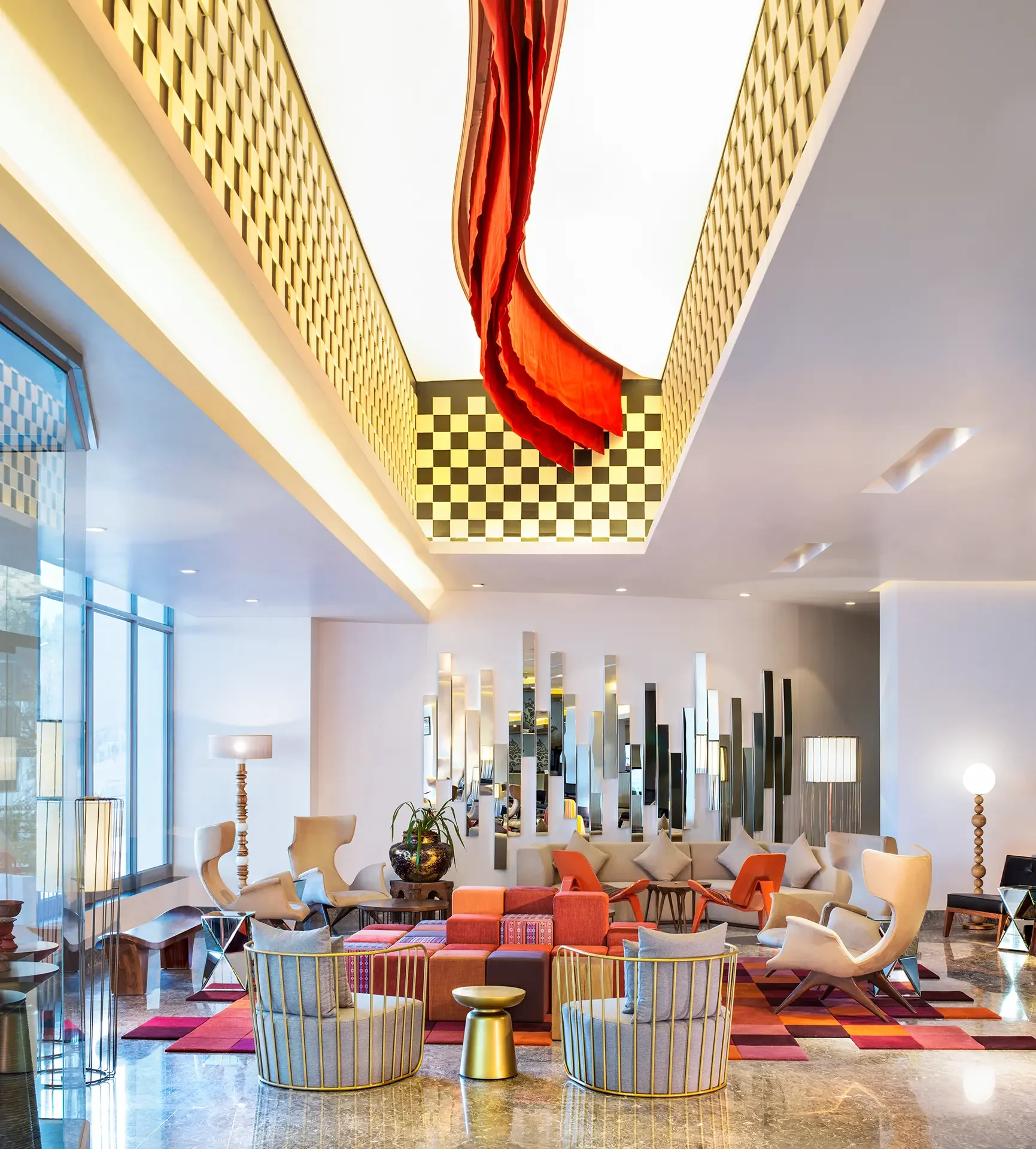 Modern hotel lobby with colorful seating, a large decorative plant, gold accent furniture, wall mirrors, and geometric carpet pattern.