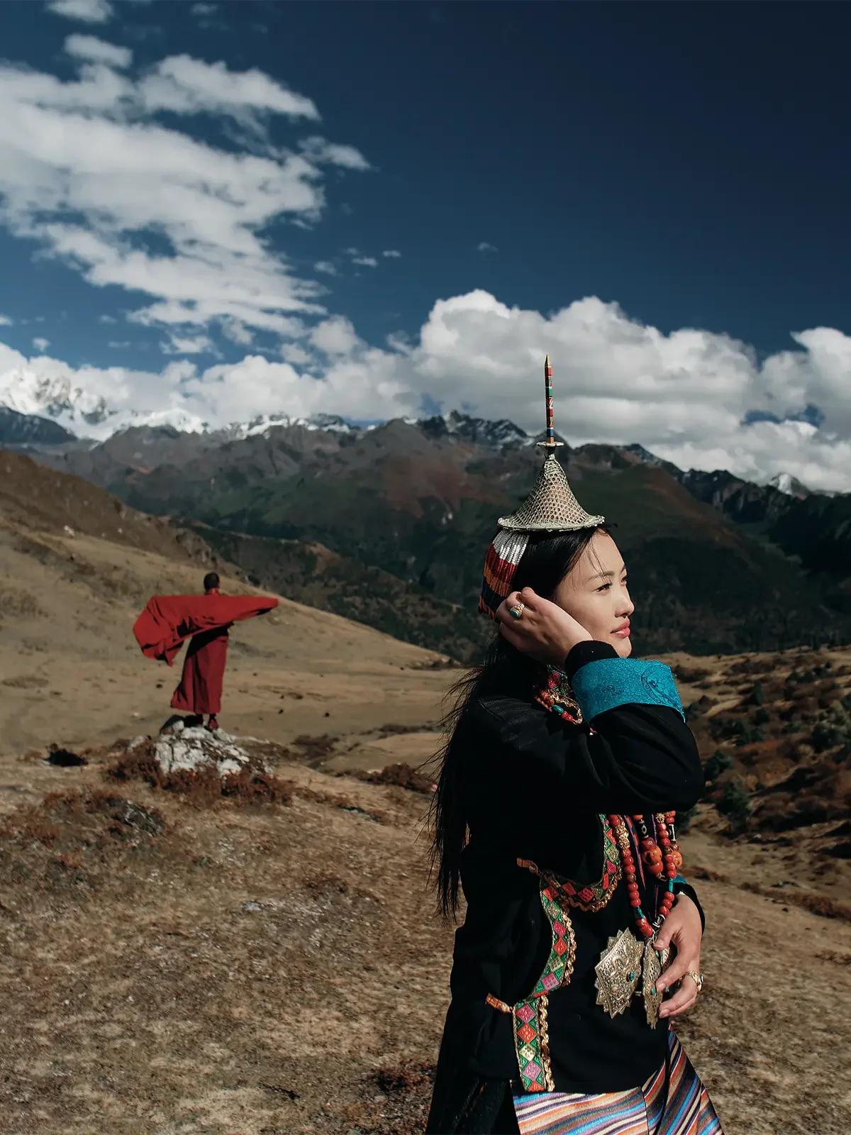 A woman in traditional attire standing outdoors with mountains and a cloudy sky in the background, while a person in red traditional clothing stands in the distance.