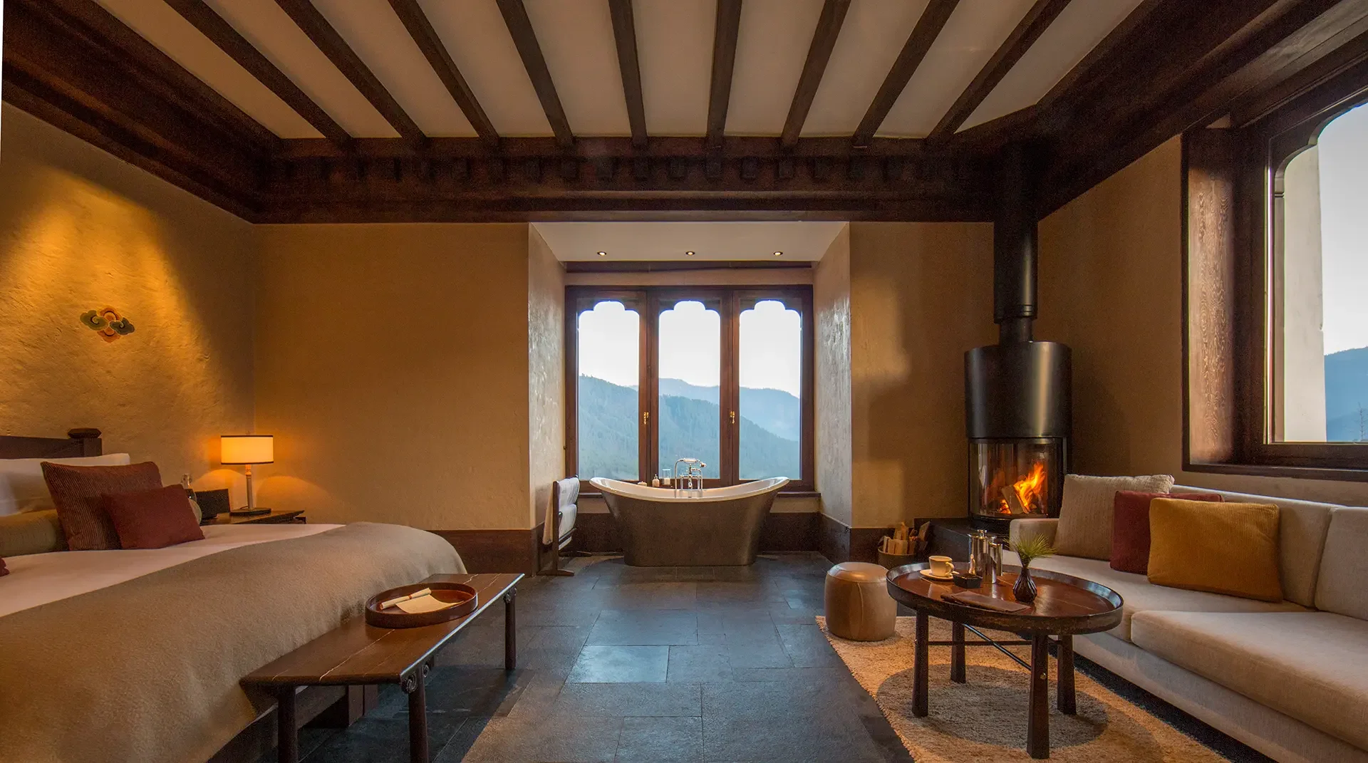 A cozy mountain cabin bedroom with a wooden ceiling, a large bed with pillows, a nightstand with a lamp, a sitting area with a couch and pillows, a fireplace with a fire, and a window showing mountain scenery.