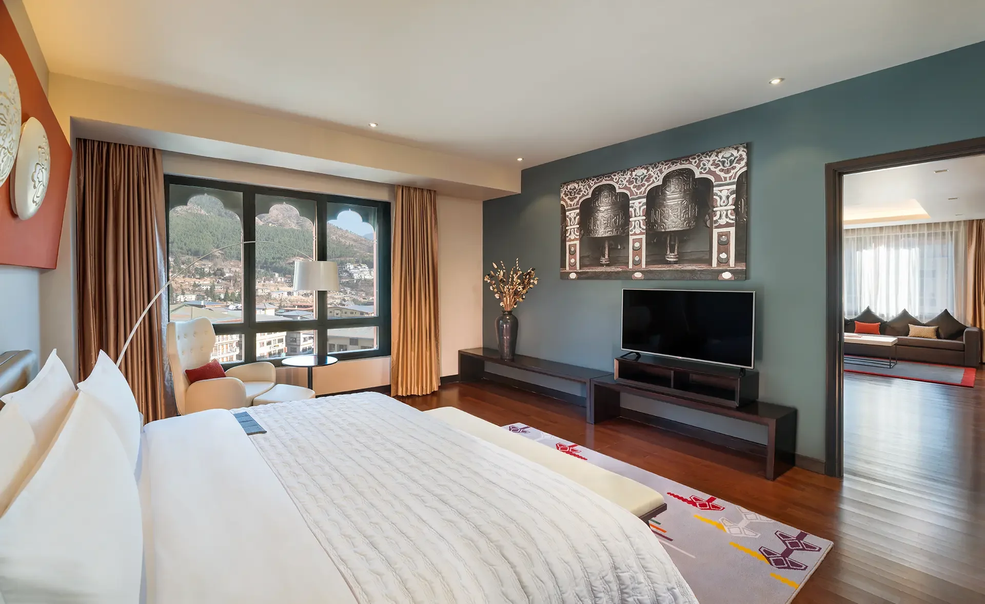 Hotel room with a large bed, white bedding, a sitting area with a white armchair and side table near a window, a flat-screen TV on a low stand, and a large wall art piece above the TV. There are curtains, vases with decorative branches, and a view of mountains through the window.