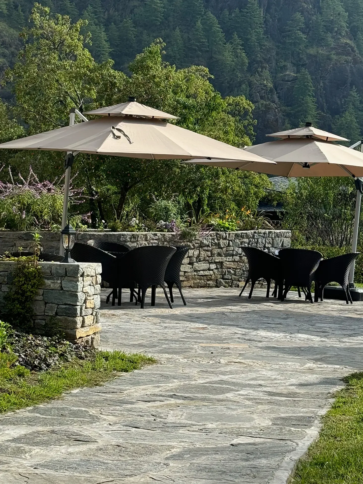 Outdoor patio area with black chairs around tables and white umbrellas, stone wall, green trees, and a mountainous background.