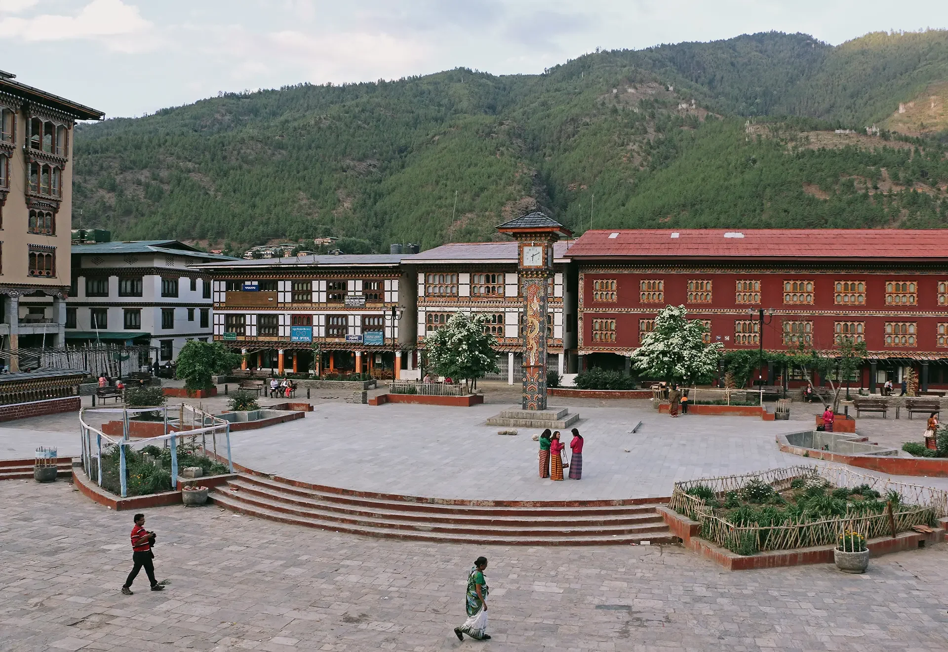 A town square with a large open paved area, a few trees, and a small raised garden bed with plants. Several people are walking or sitting, including a group of three women wearing traditional clothing. There are buildings with traditional architectural details, and a mountain in the background.