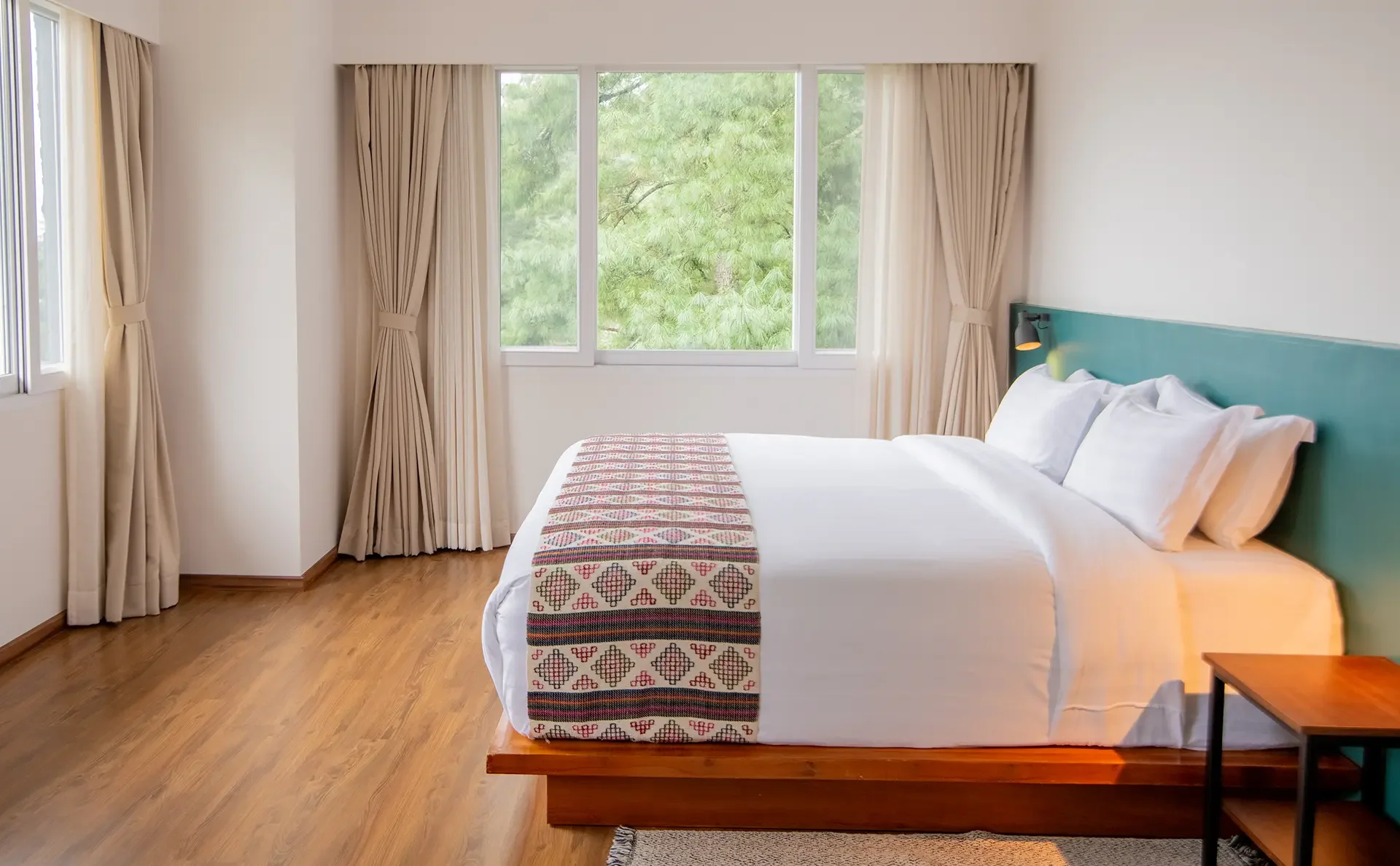 A cozy bedroom with a large bed covered in white linens and pillows. The bed has a embroidered runner at the foot, and a teal headboard with a reading lamp attached. There are beige curtains on the windows, and a nightstand on the right. Outside the windows, green trees can be seen, letting natural light into the room. The floor is wooden, and a small rug is partially visible.