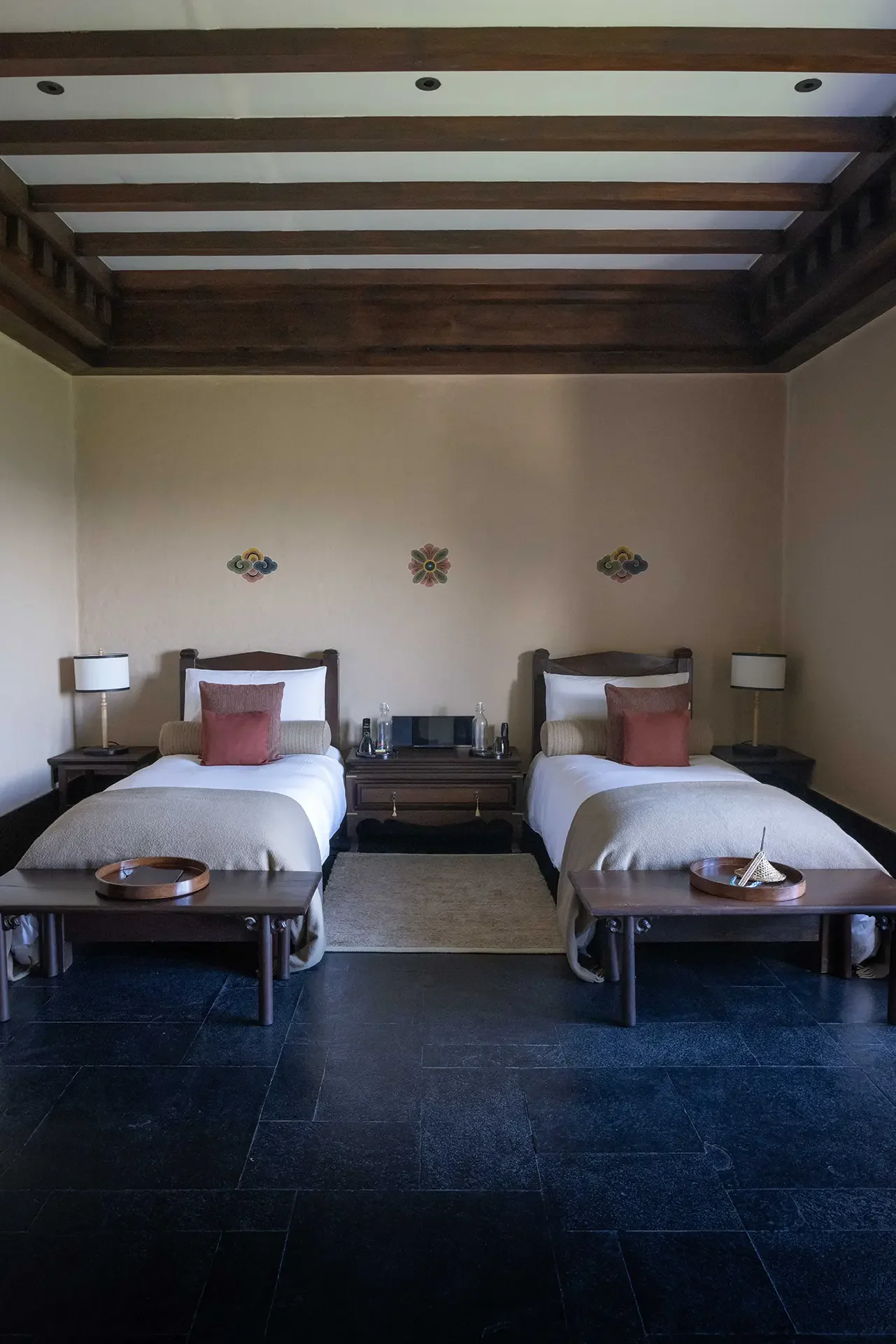 Hotel room with two beds, nightstands, lamps, and decorative wall hangings, featuring a dark wood ceiling with white beams.