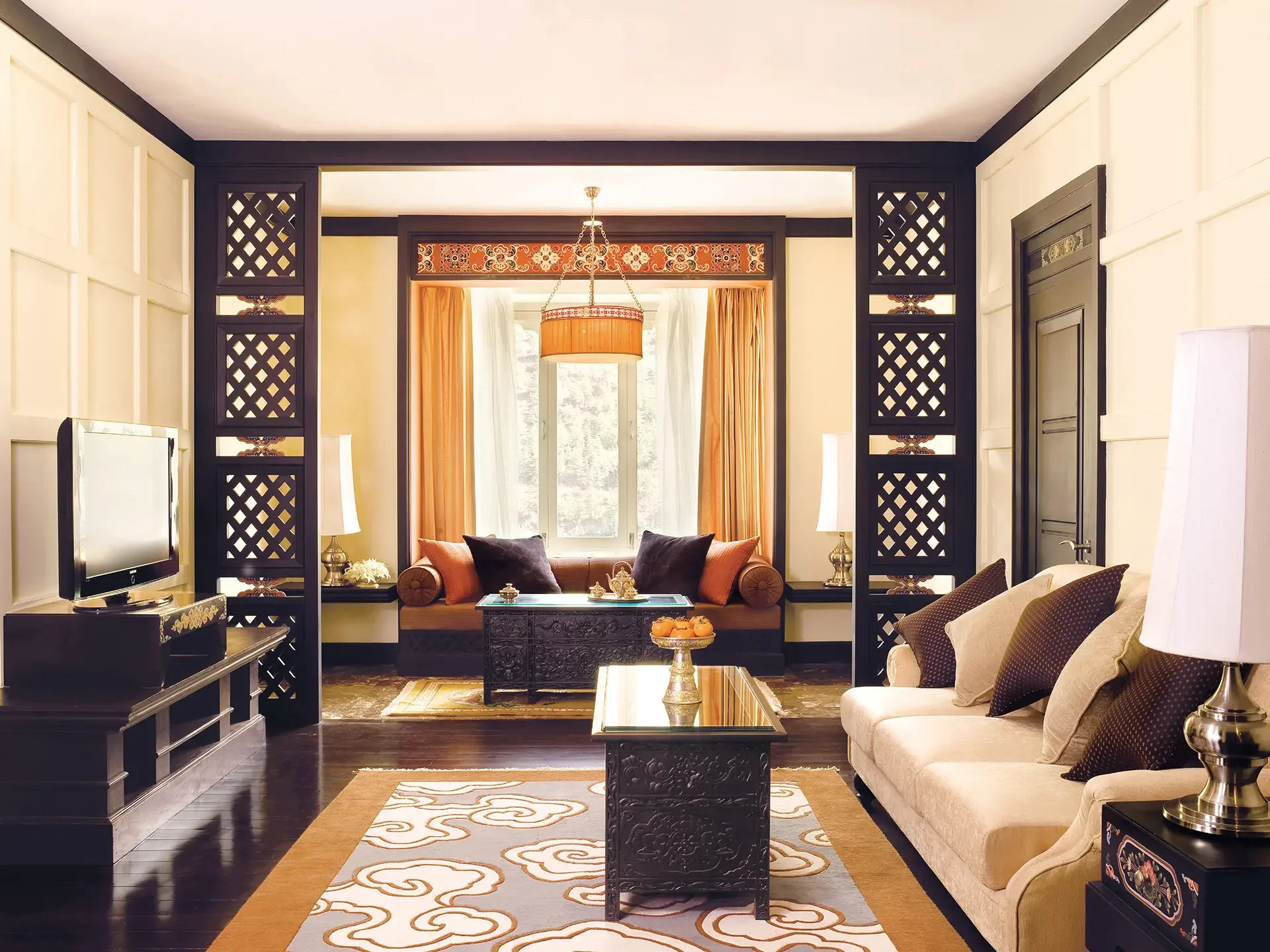 A living room with cream sofas, brown and orange cushions, black ornate coffee tables, a patterned rug, and large windows with orange curtains, decorated in a luxurious style.