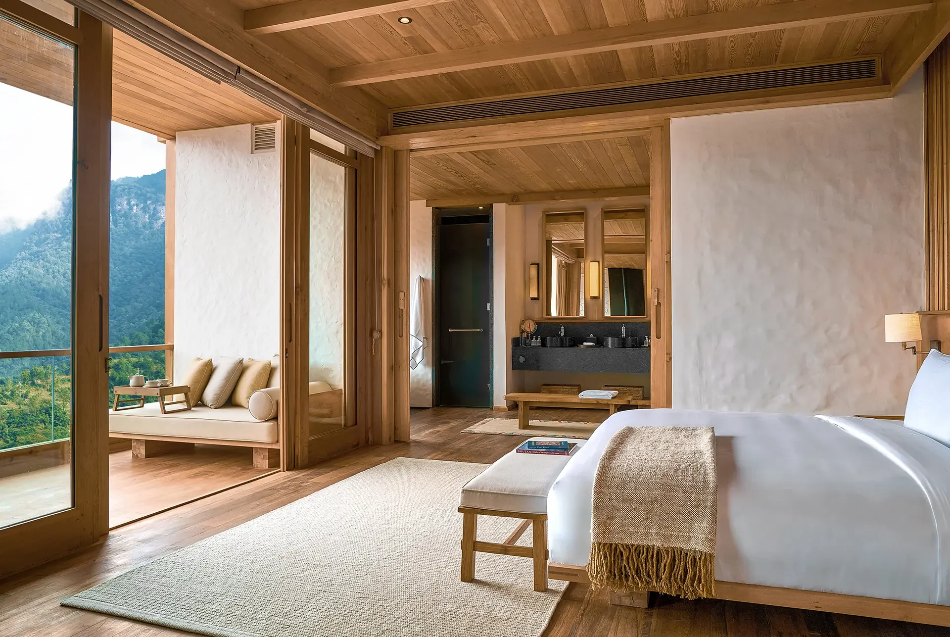 A luxurious bedroom with wooden ceiling and walls, featuring a large bed with white bedding and a beige throw blanket, a small wooden bench, a magazine on the floor, a seating area with a cream-colored sofa and cushions outside the bedroom, and large sliding glass doors overlooking a mountain landscape.