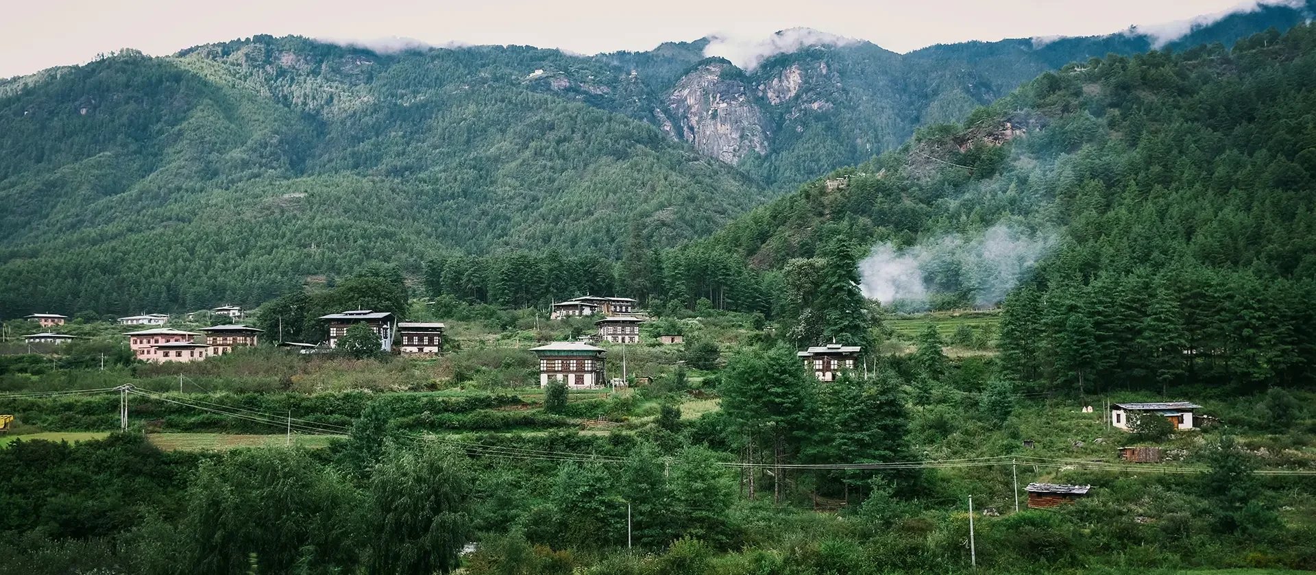 Mountains with dense green forests and a small village with traditional houses at the hillside, some smoke rising from the trees.