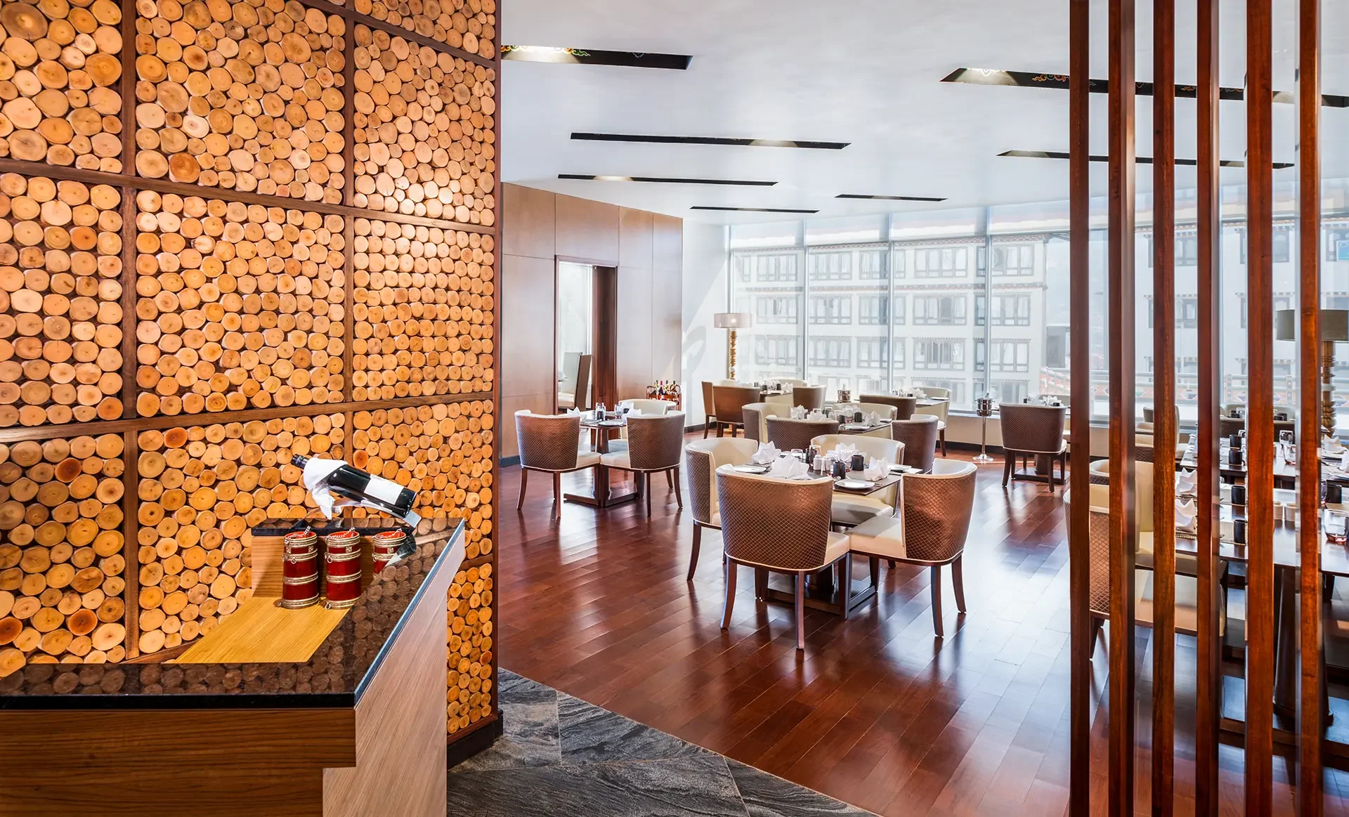 Elegant restaurant dining area with white tablecloths, chairs, and floor-to-ceiling windows showing a cityscape outside. A wall of logs is on the left, and wooden-slatted dividers are on the right.