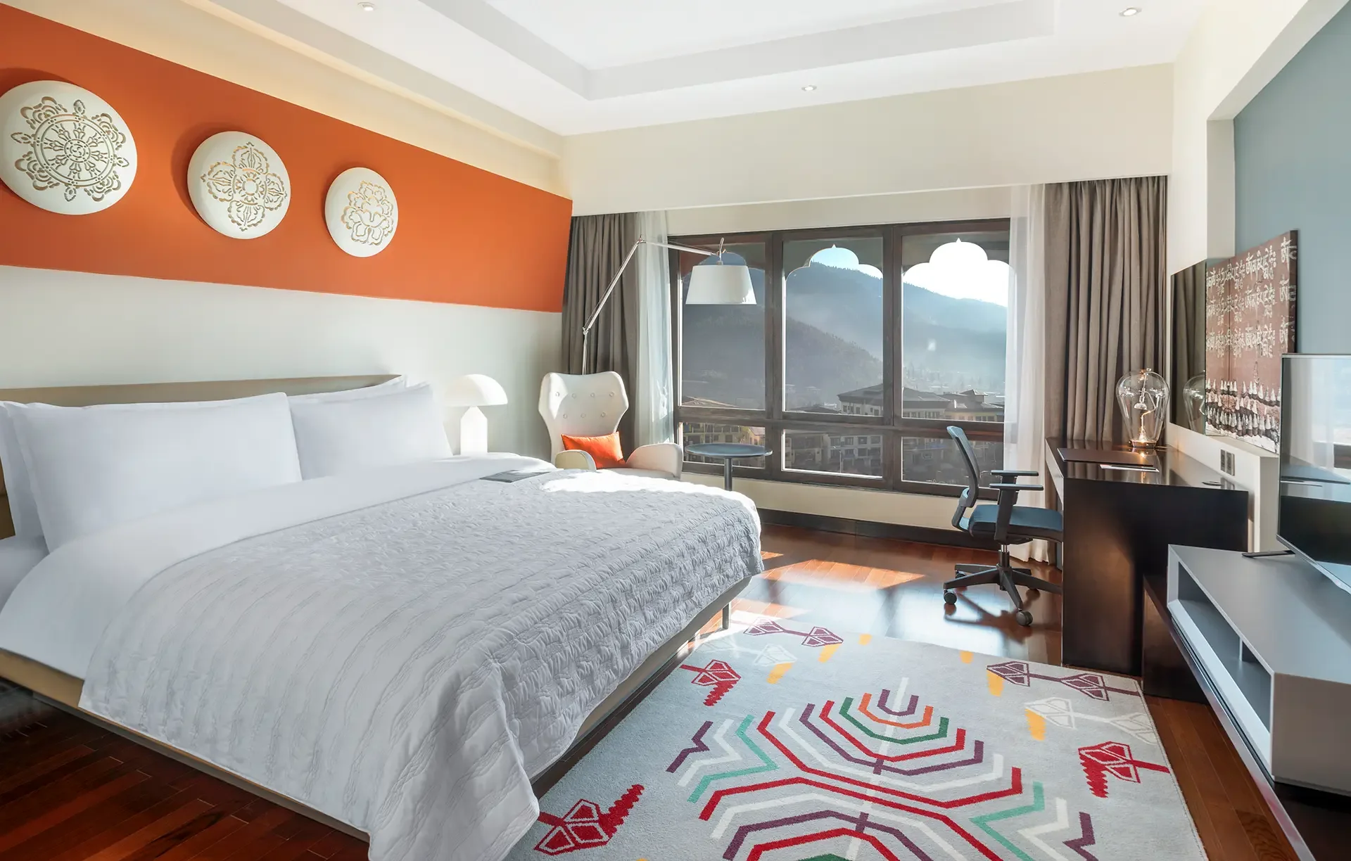Modern hotel room with large bed, colorful geometric area rug, and a view of mountains outside the window.