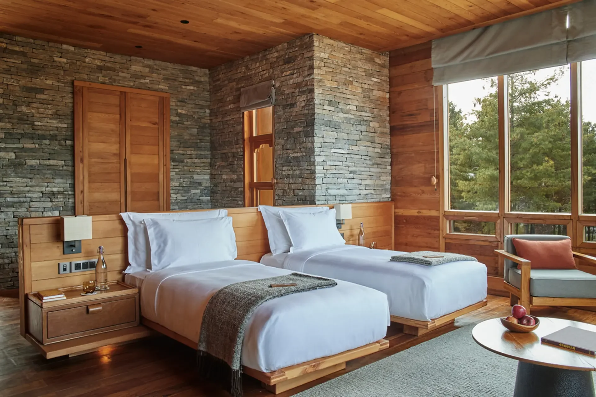 A bedroom with two beds with white bedding, wooden headboards, and bedside tables; a stone wall and large window with greenery outside.