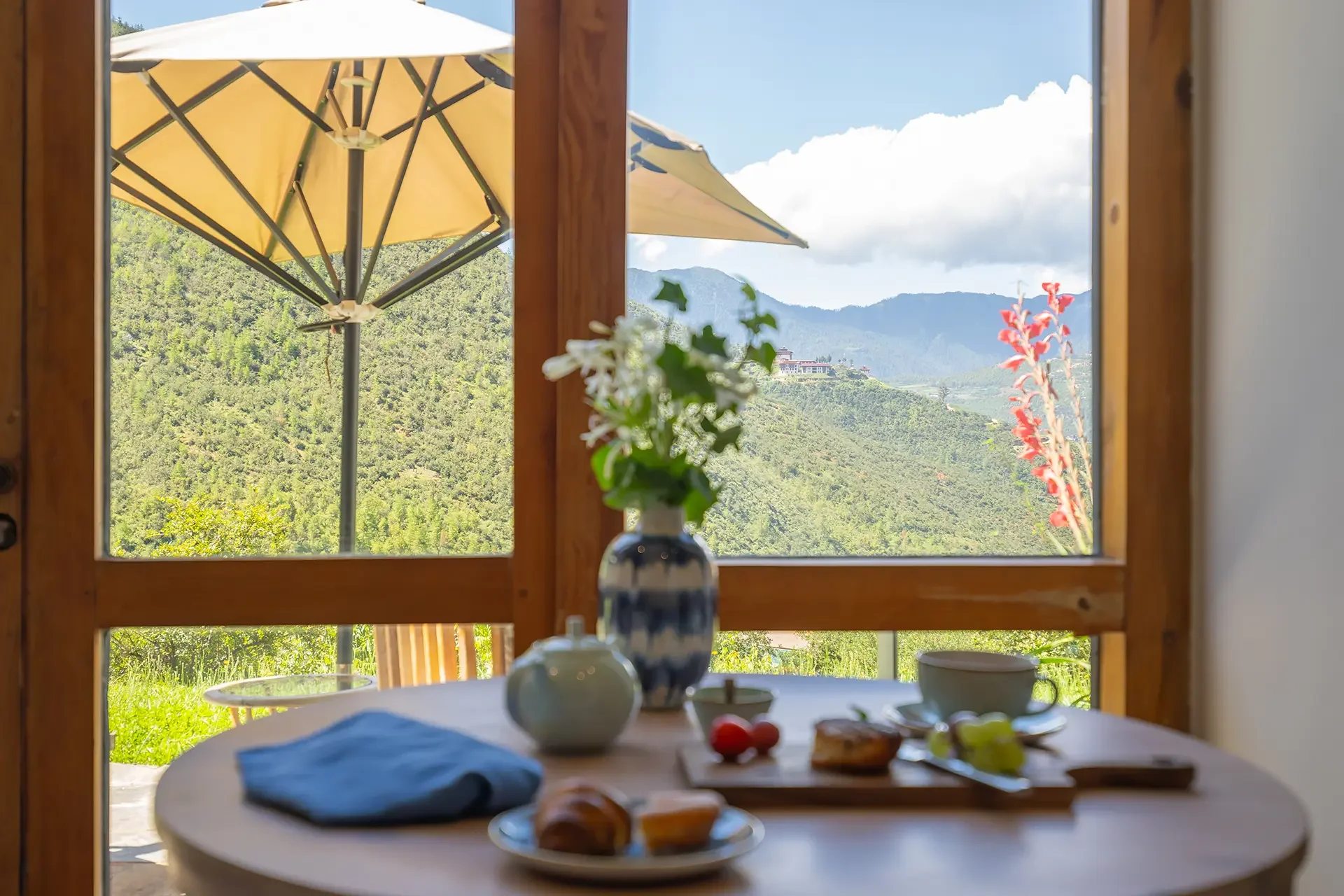 View through a window showing a breakfast table with bread and fruit, a vase with white flowers, and an outdoor scene with a yellow umbrella, green mountains, and partly cloudy sky.