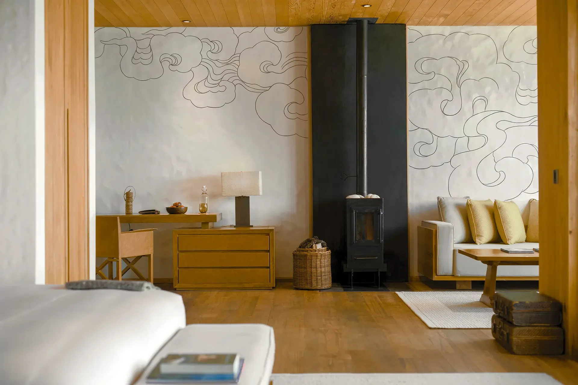 Living room with wooden ceiling, a black stove, a white sofa with yellow pillows, a wooden coffee table, a white wall with a black line drawing of clouds, and a small desk with a lamp and decorative items.