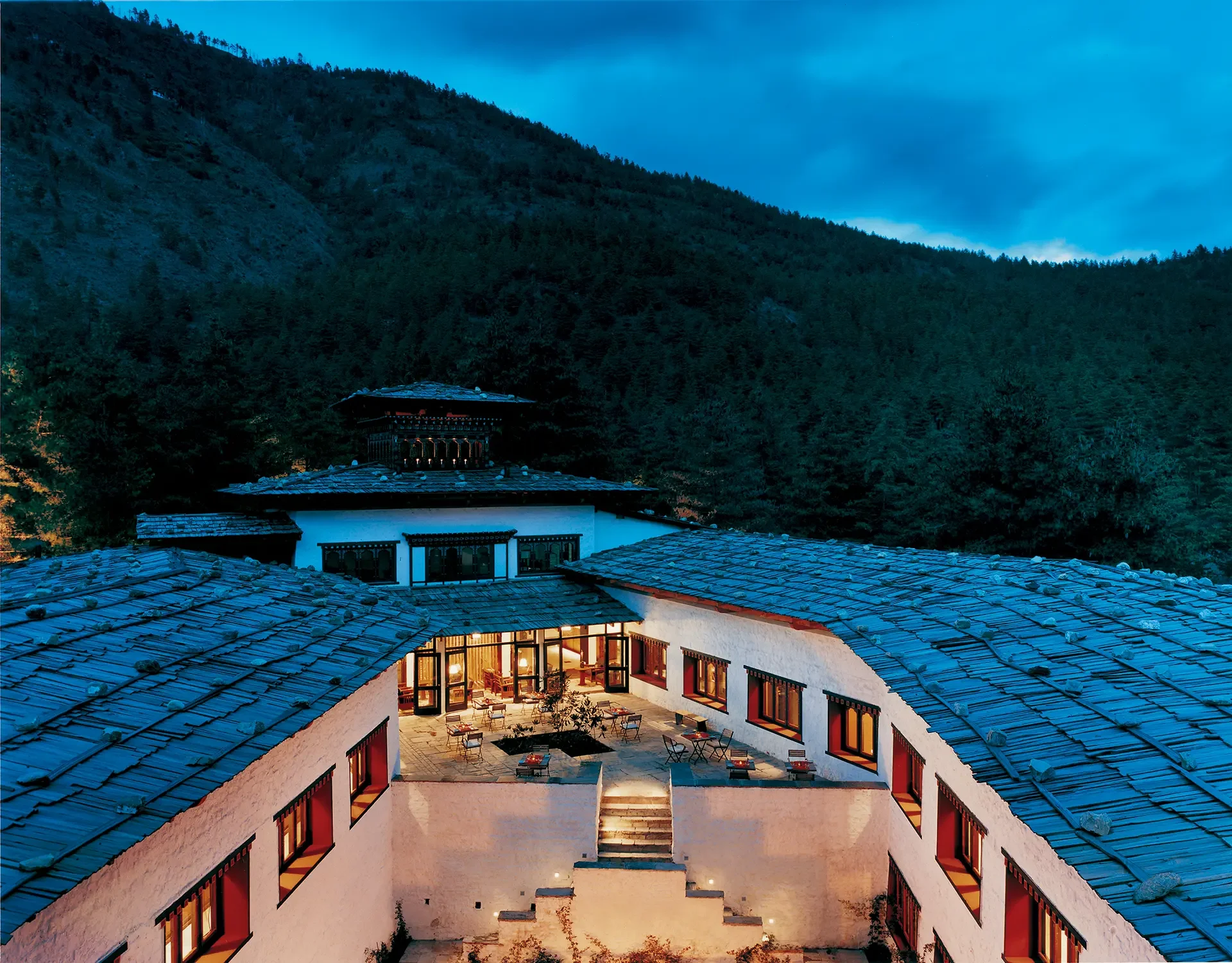 A view of a mountain resort at dusk with a courtyard, tables, and chairs, surrounded by white buildings with dark window frames and tiled roofs, and a pagoda style roof in the background.