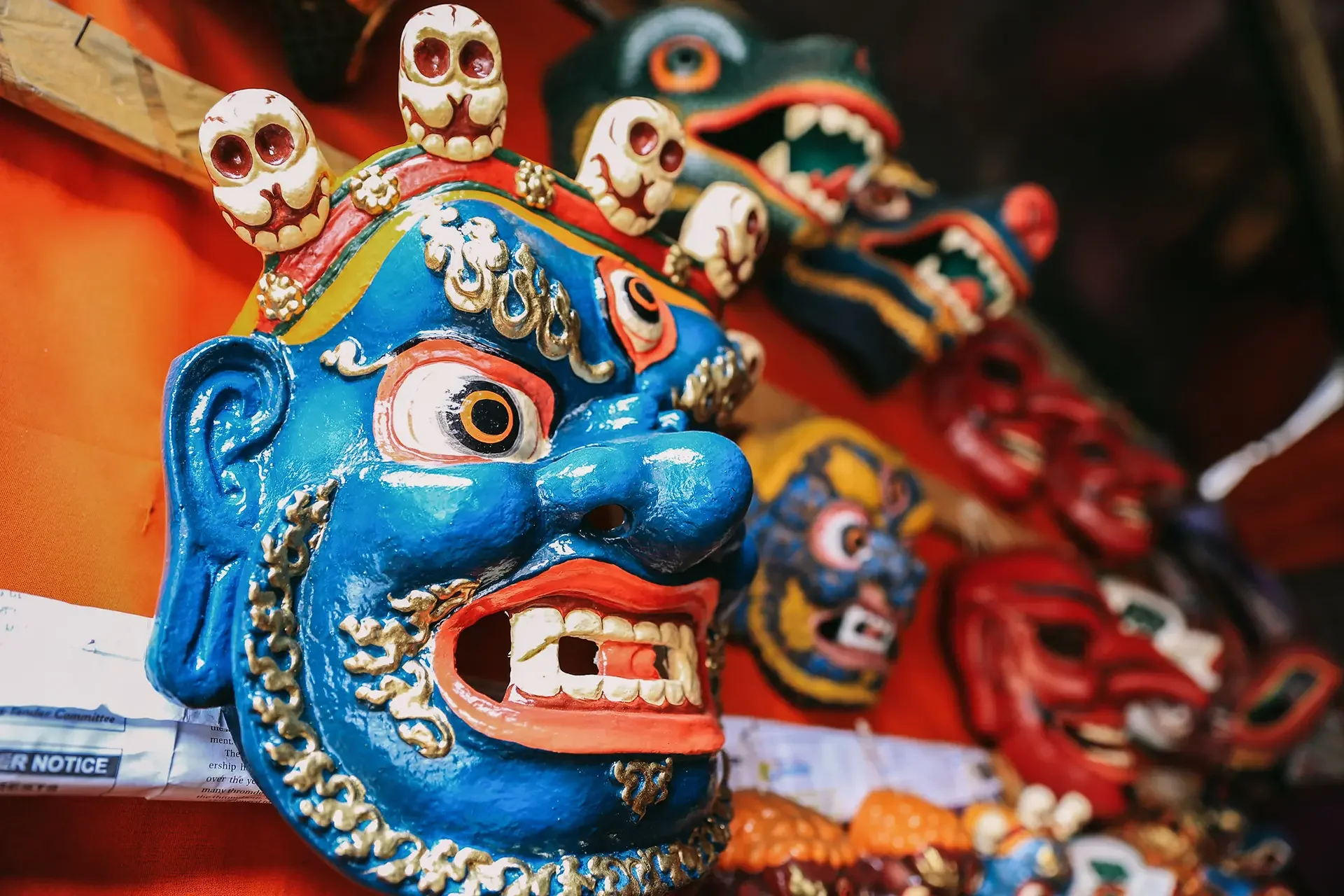Colorful traditional masks displayed on a red surface, featuring a blue face with gold accents and skull motifs, along with others in red, black, and multicolored designs.