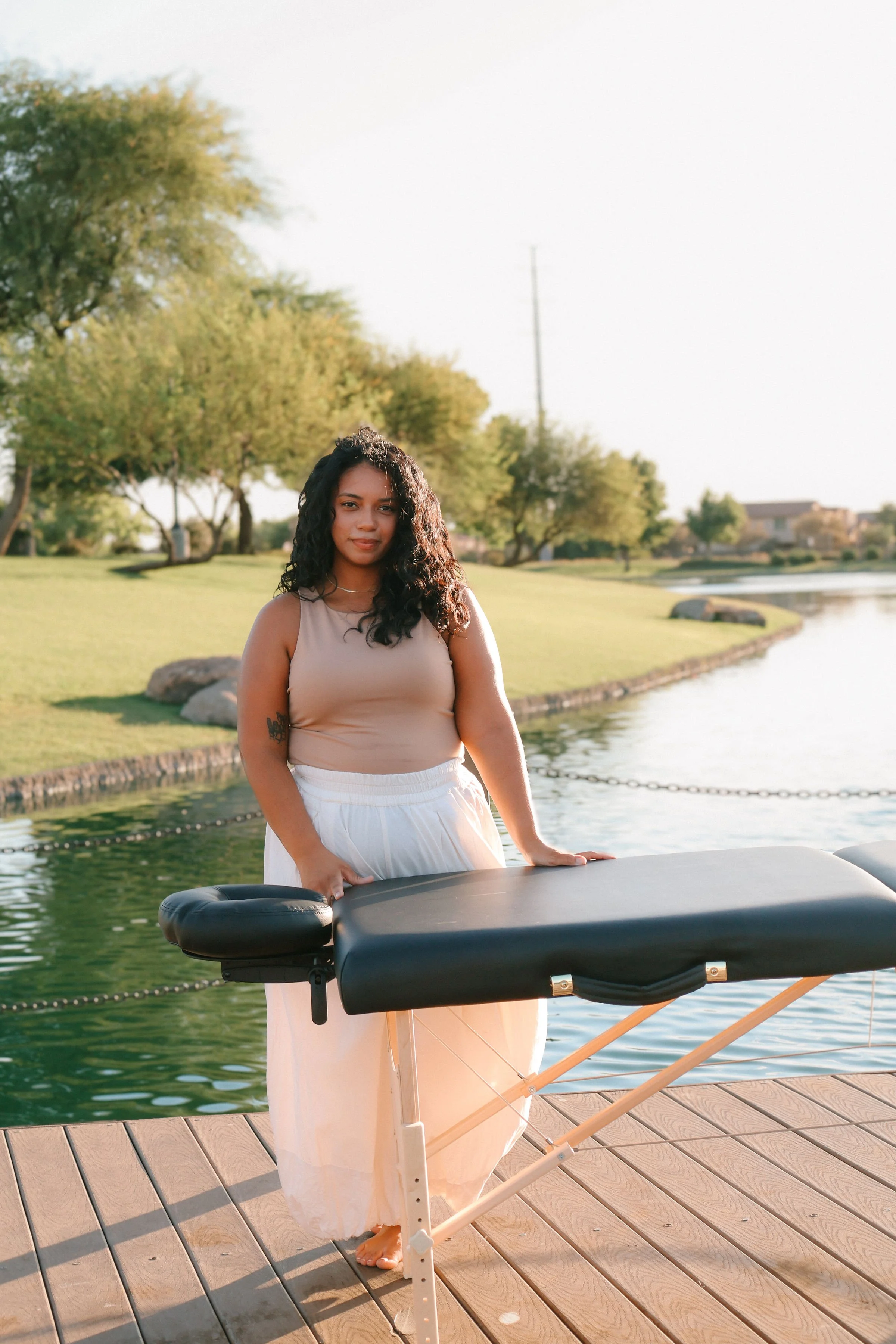 A woman with curly black hair stands on a wooden dock, leaning on a massage table, with a lake, green trees, and houses in the background.