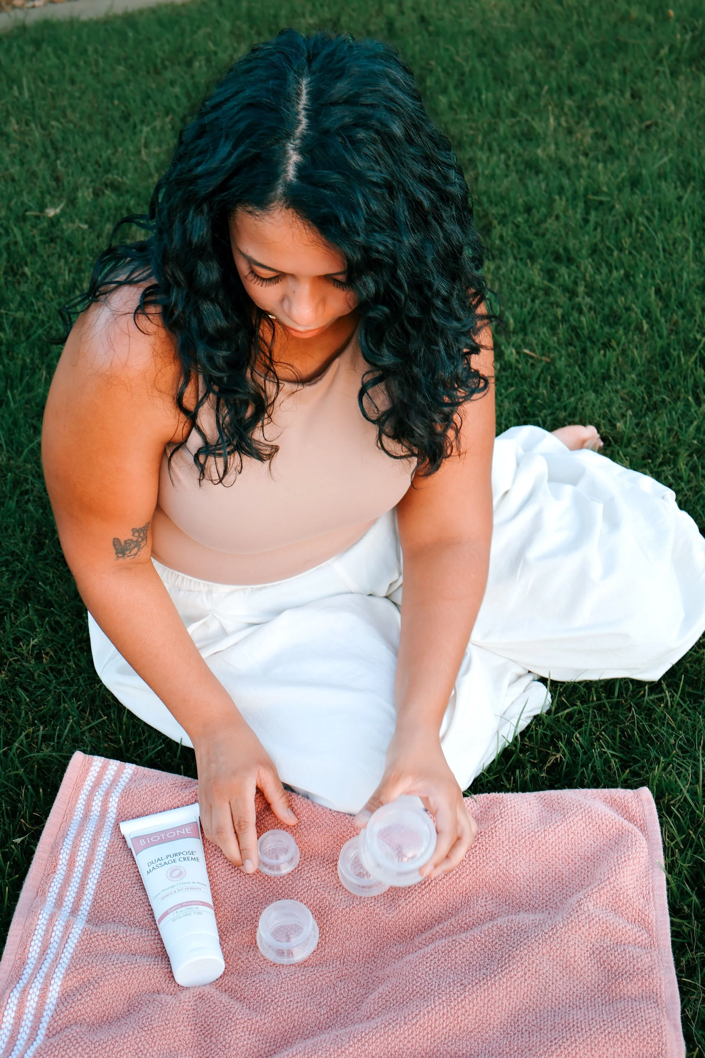 A woman with dark, curly hair, wearing a beige top and white pants, sitting on green grass outdoors, surrounded by skincare products and a pink towel, engaged in a skincare routine.
