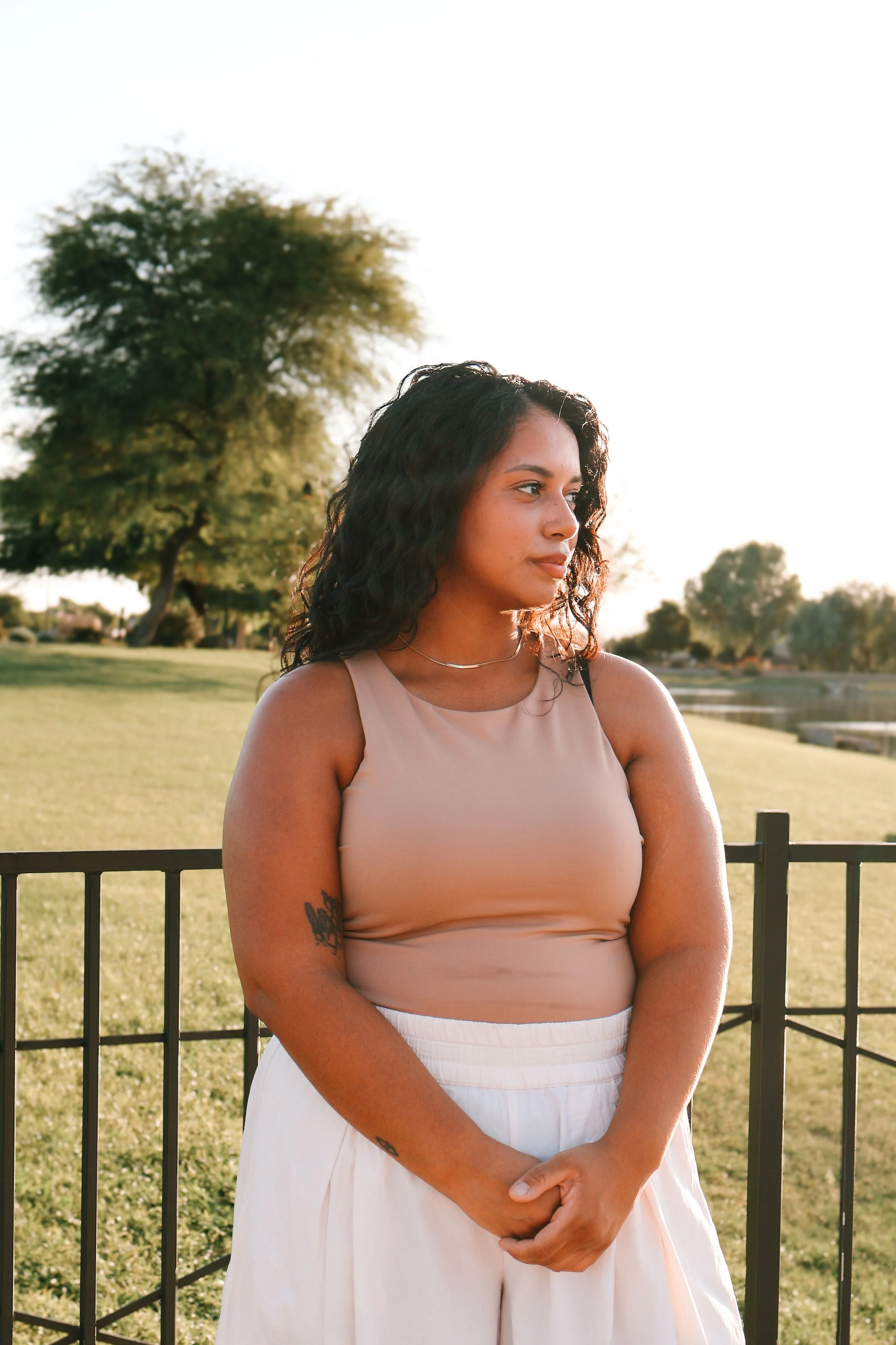 A young woman with curly dark hair, wearing a beige sleeveless top and white skirt, stands outdoors near a black fence with a park and trees in the background, during late afternoon sunlight.