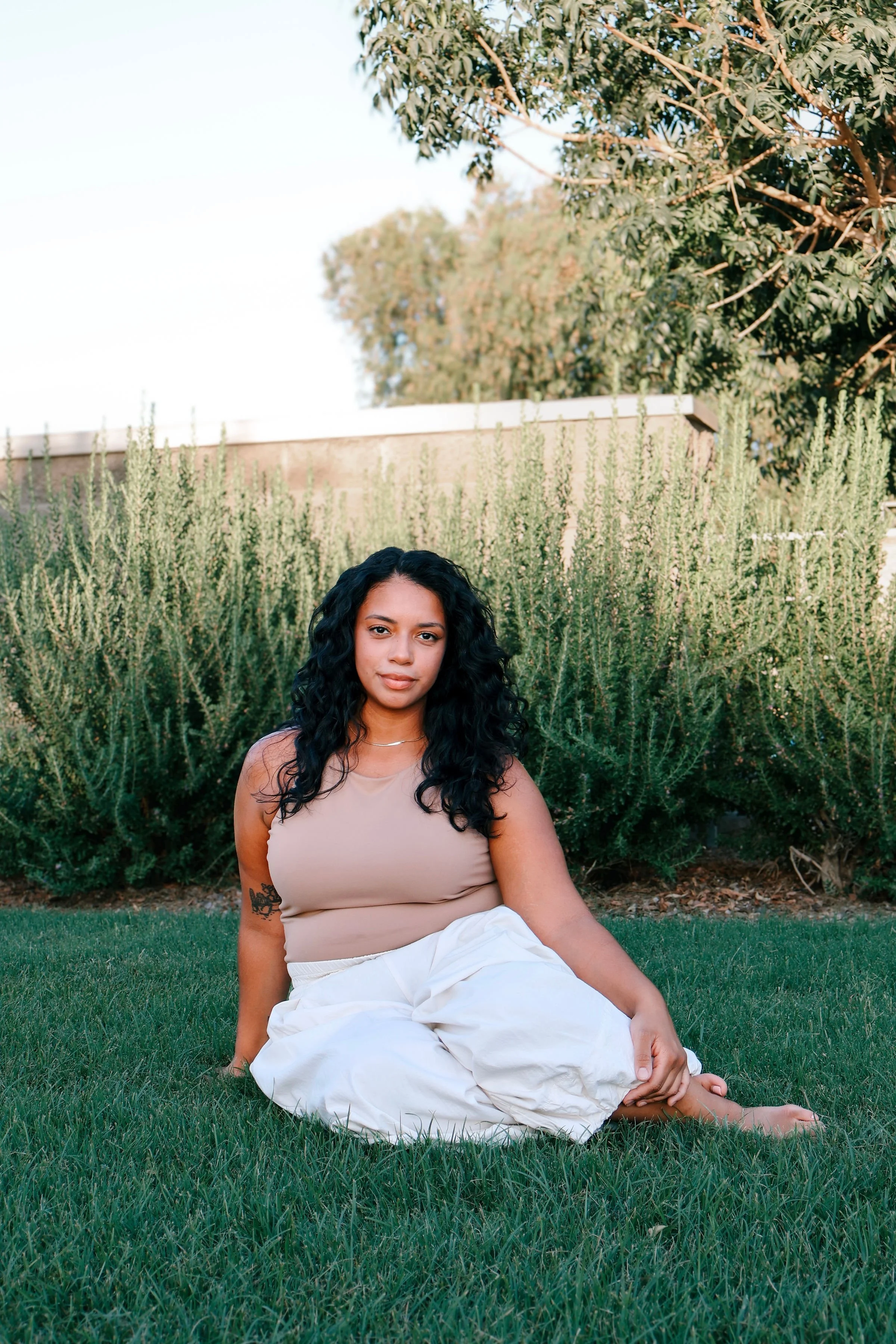 A woman with long curly black hair, sitting on the grass in a garden, wearing a beige sleeveless top and white pants, with a tattoo on her arm, surrounded by green plants and trees.