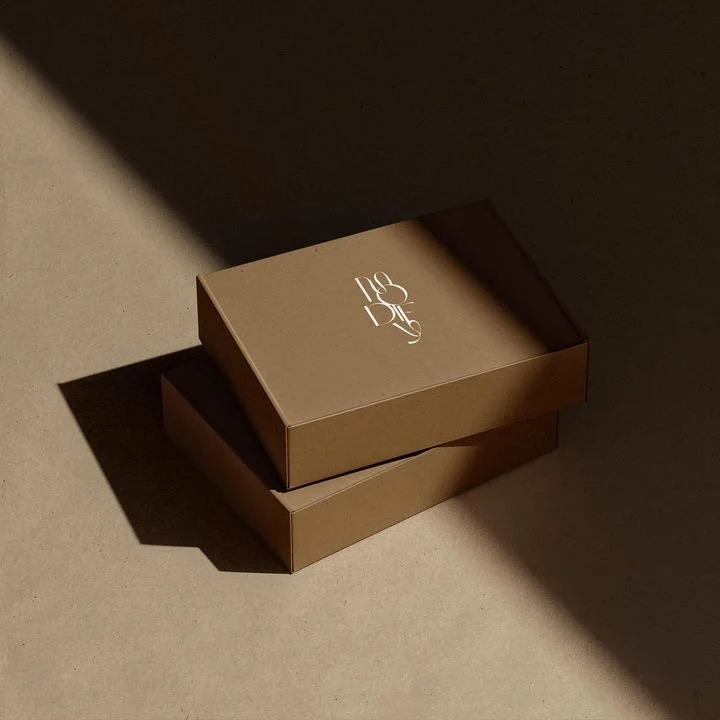 Two brown cardboard boxes stacked on top of each other with a white logo on the lid, placed on a beige surface with shadows.