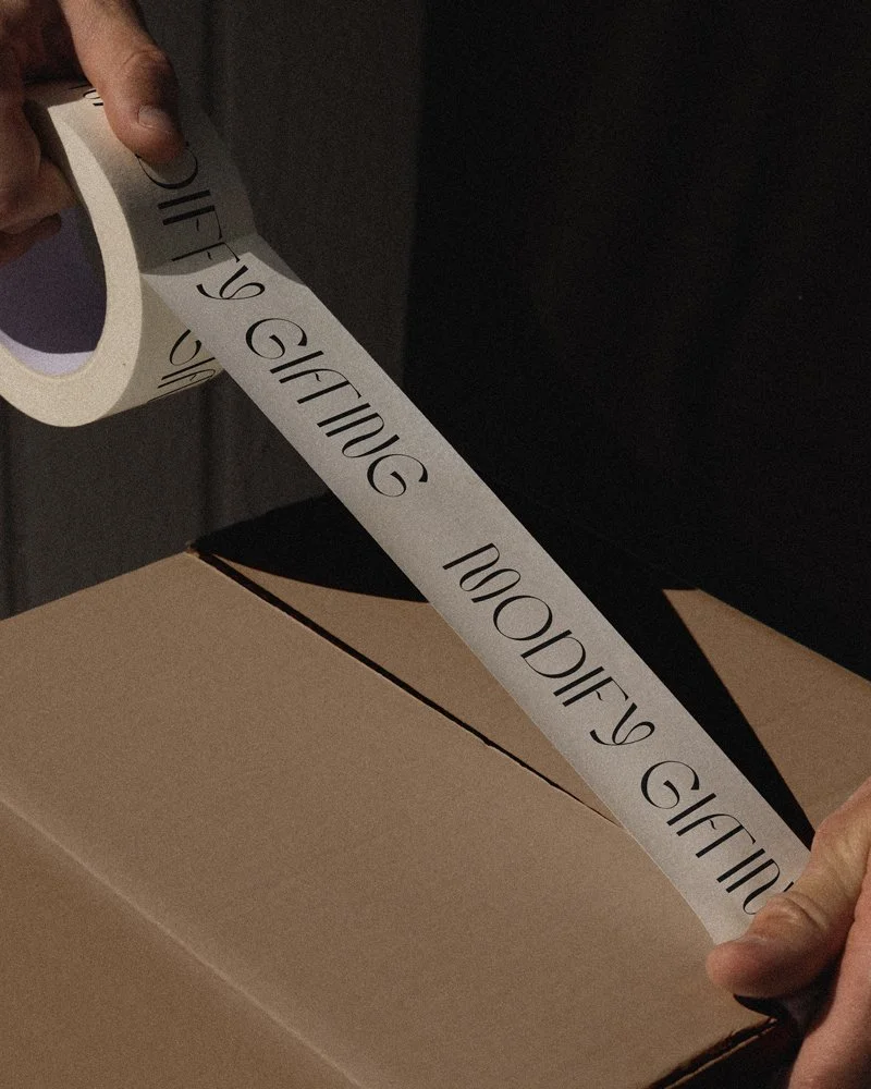 Person applying packing tape with the words 'GIFT' to a cardboard box.