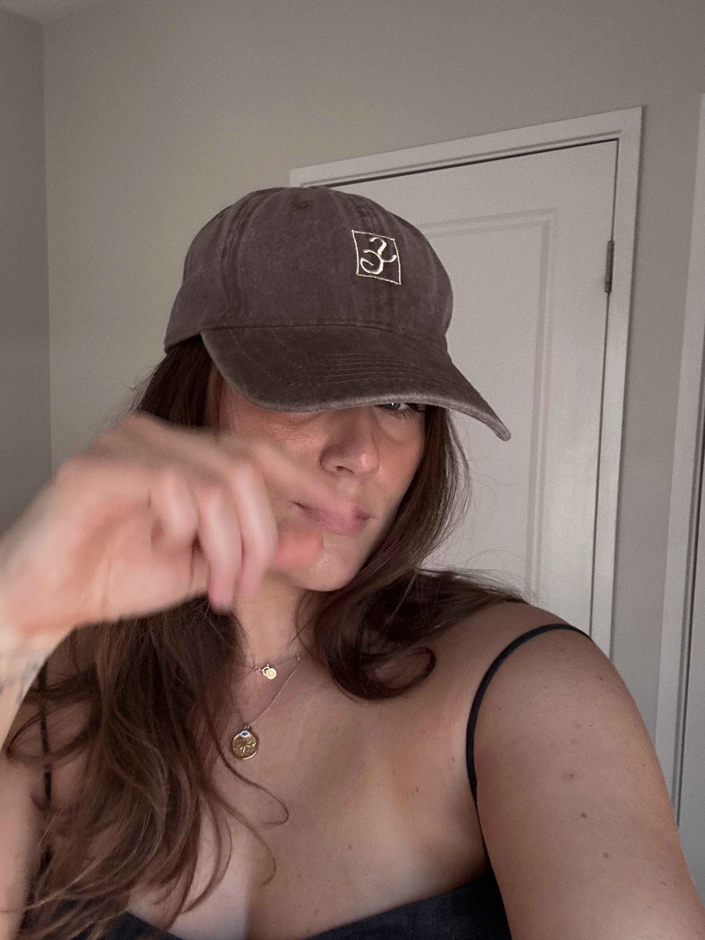A woman with long brown hair wearing a gray cap with the number 23 embroidered on it, a black strap top, and two gold necklaces, standing in front of a white door in a room.