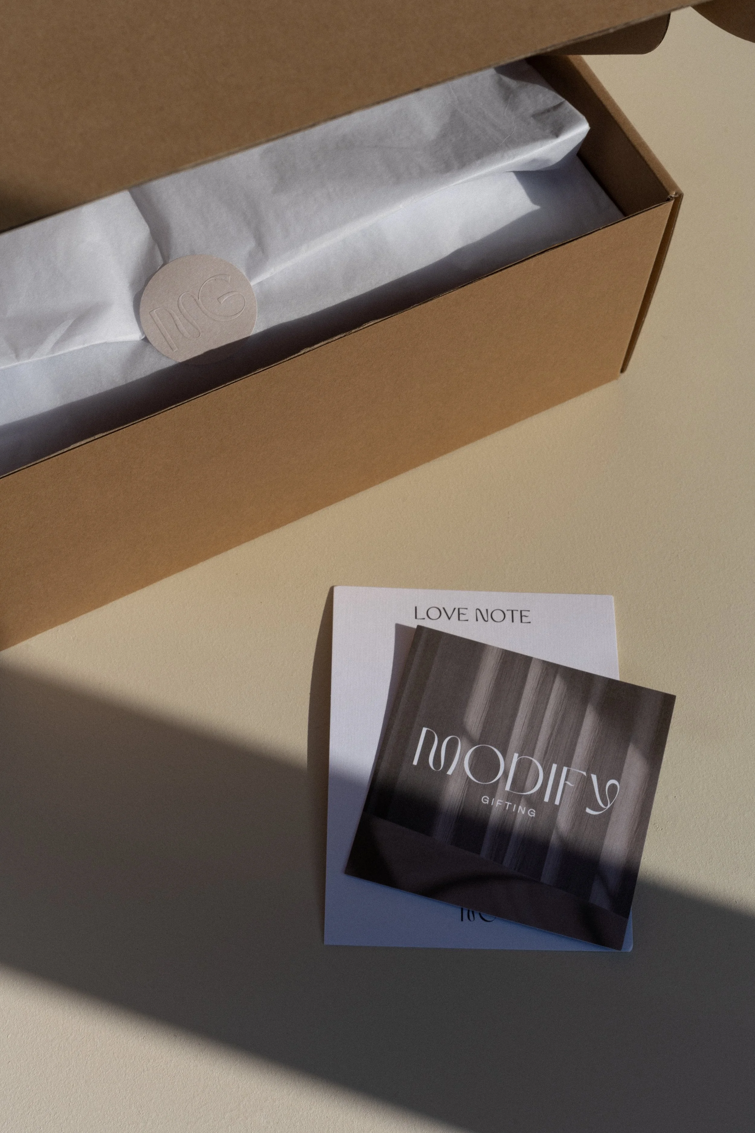 Open cardboard gift box with white tissue paper and a circular tag inside, next to folded note cards labeled 'LOVE NOTE' and 'MODIFY GIFTING.'