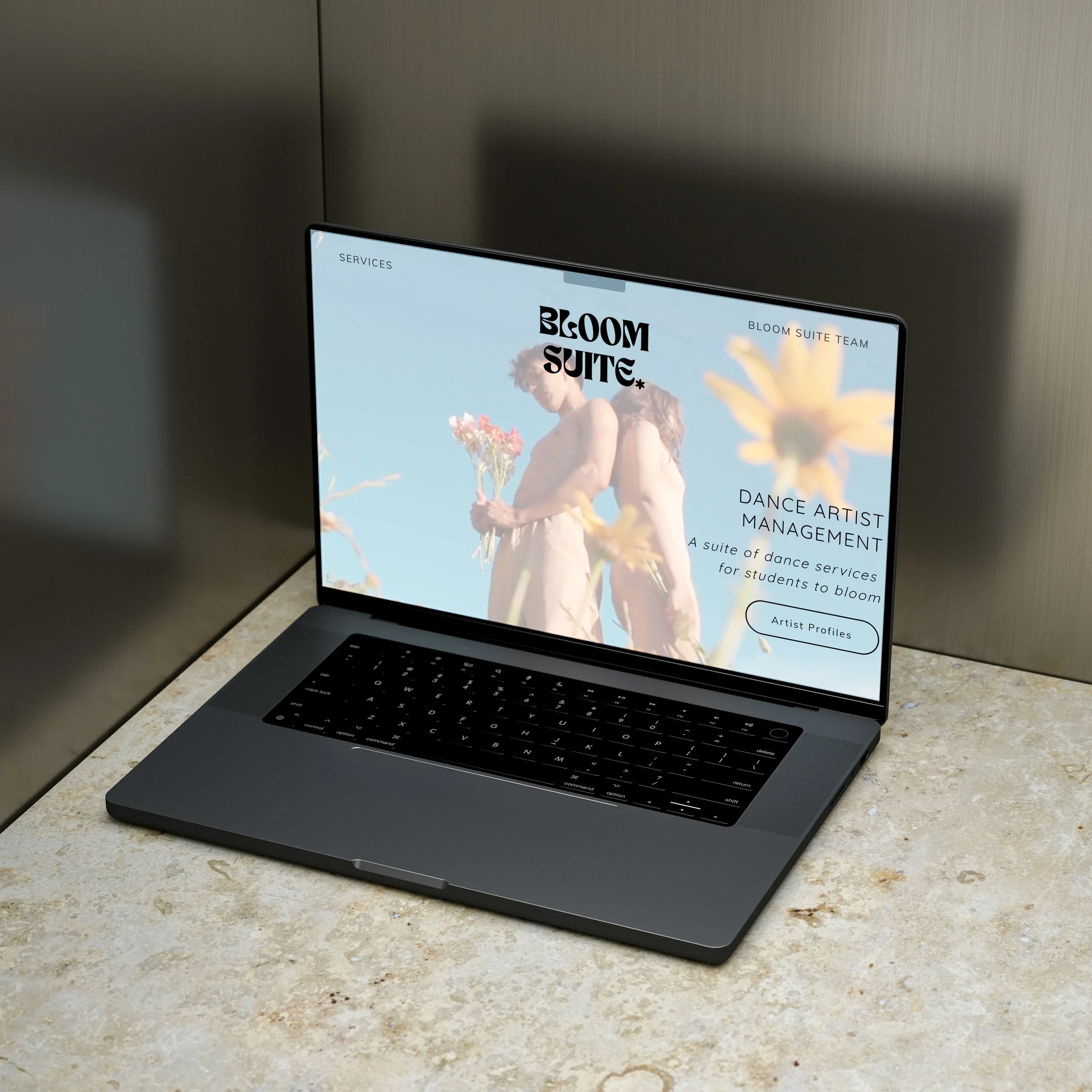 A laptop on a marble countertop displaying a website for Bloom Suite, showing two dancers holding flowers against a blue sky background.
