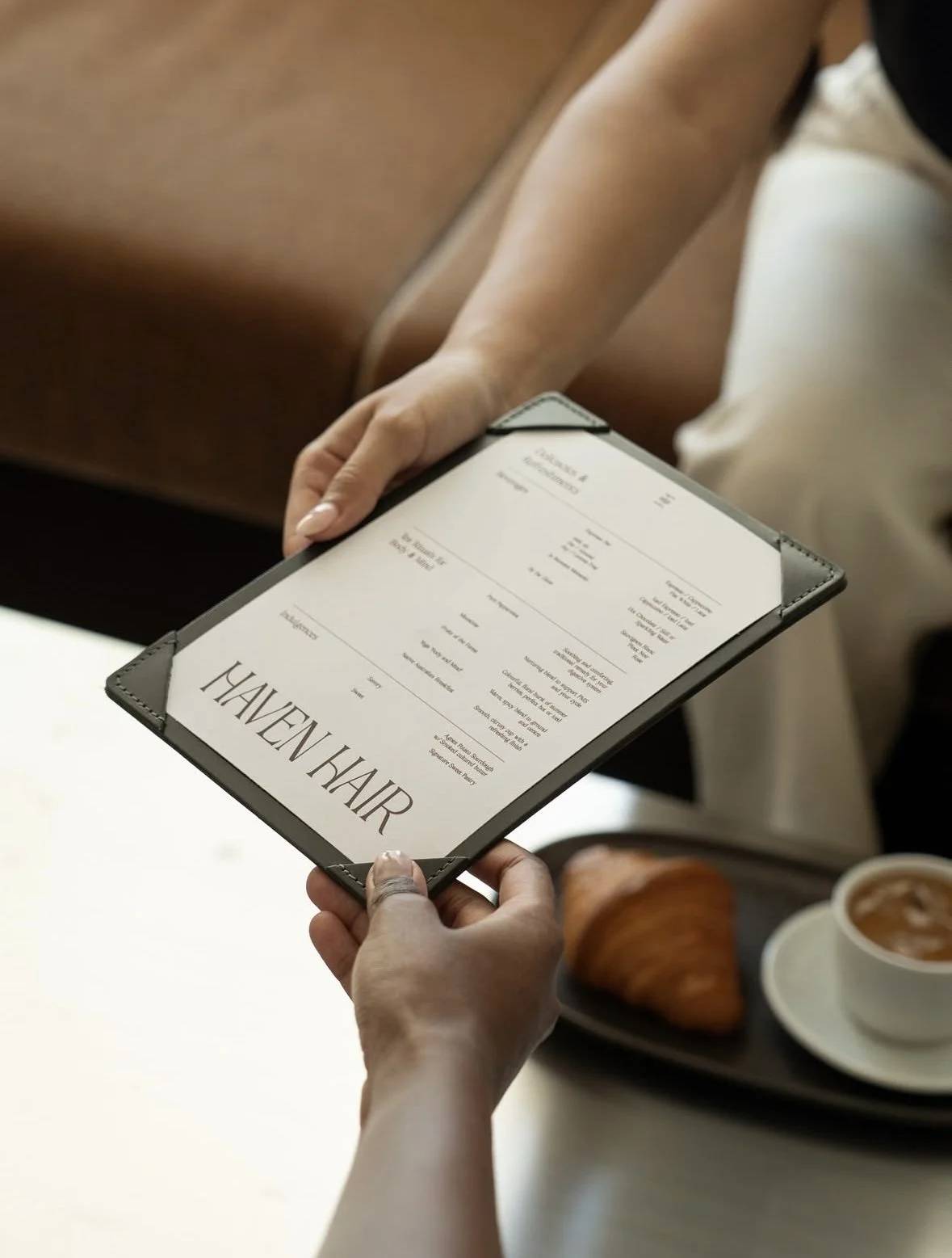 Person holding a menu with the text 'HAVEN HAIR', alongside a tray with a croissant and a cup of coffee.