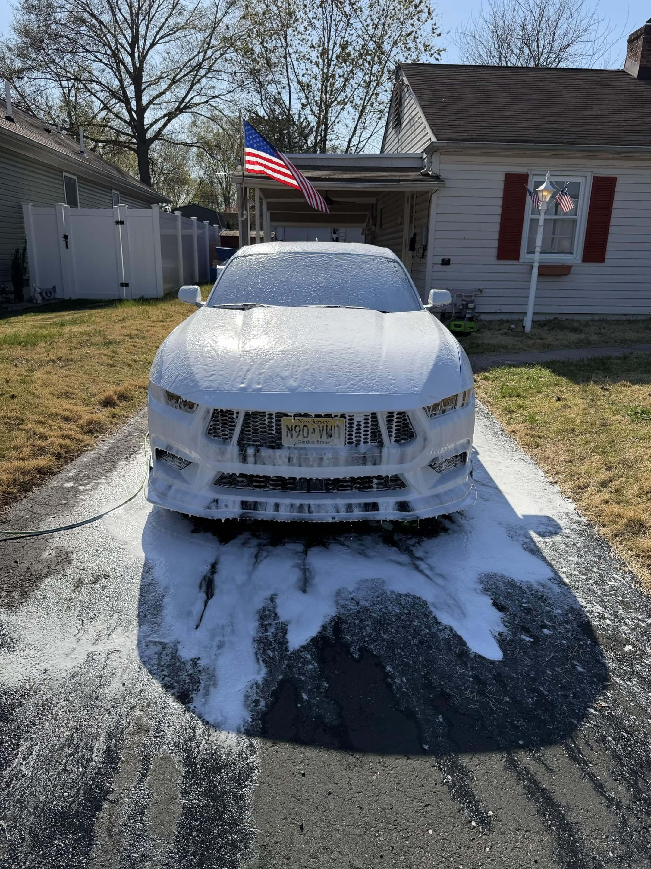 A white car is being washed with soap foam on the hood, front bumper, and windshield in a residential driveway. An American flag is on a pole in the porch area of a house with white siding and red shutters. There is a lamppost with small American flags, a white fence, and leafless trees in the background.
