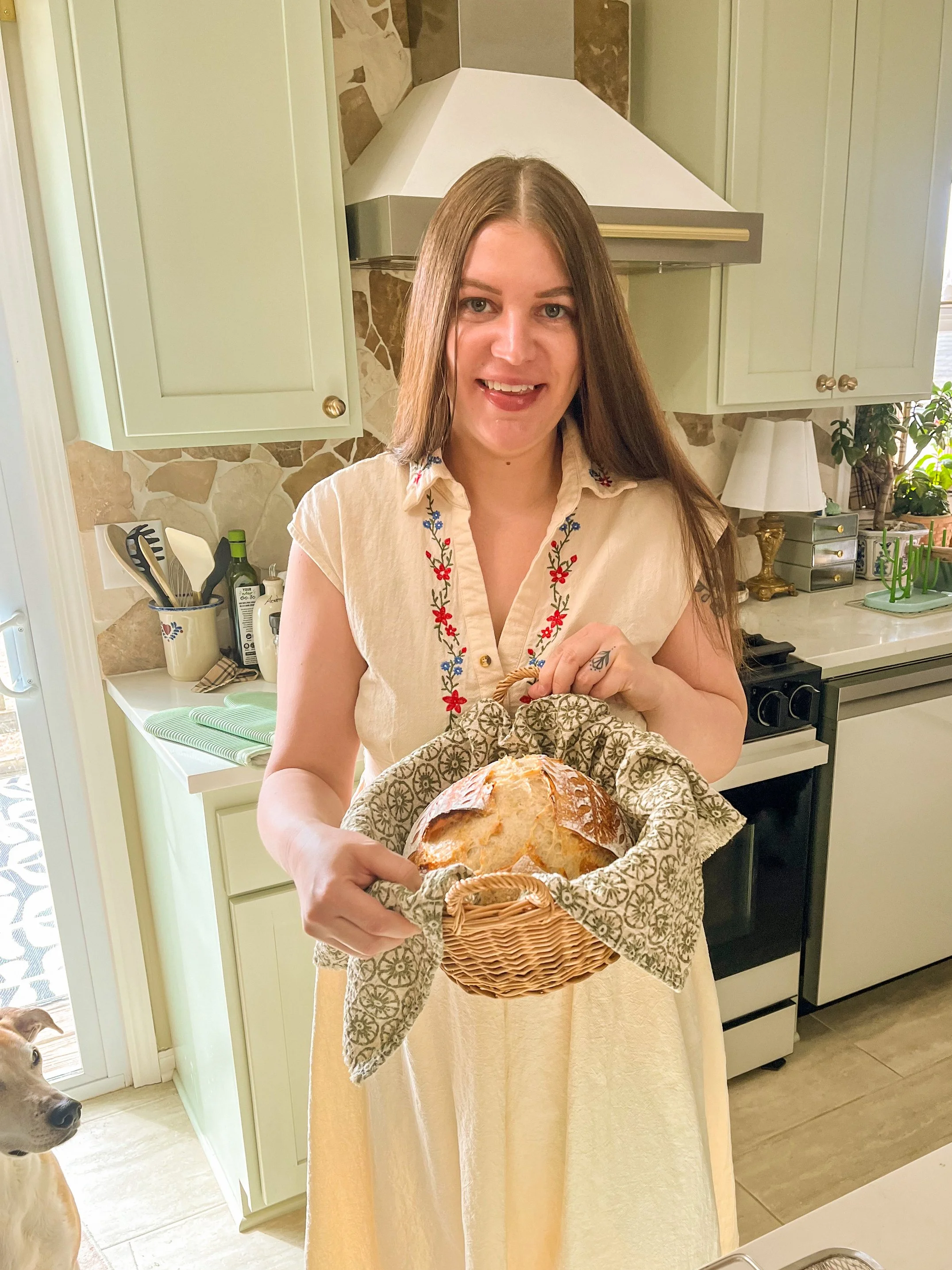 A woman with long brown hair wearing a cream-colored dress with embroidered flowers, holding a basket with a loaf of bread, standing in a kitchen with green cabinets and a stone backsplash.