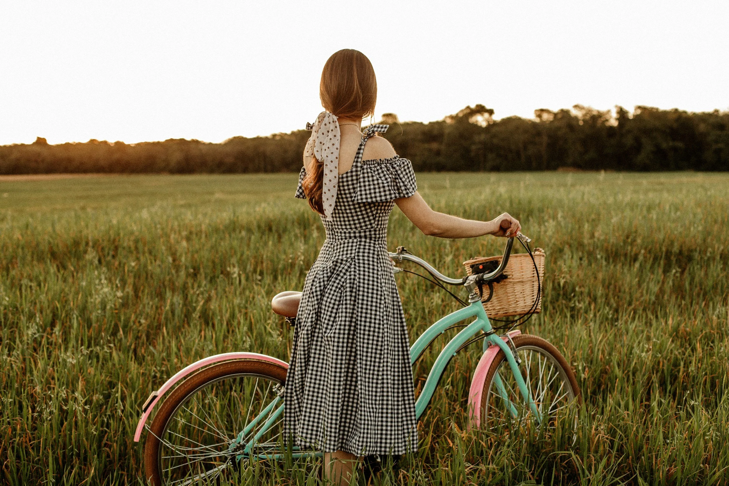A woman with long reddish hair standing in a grassy field at sunset holding a bicycle with a wicker basket in the front and gazing into the distance.