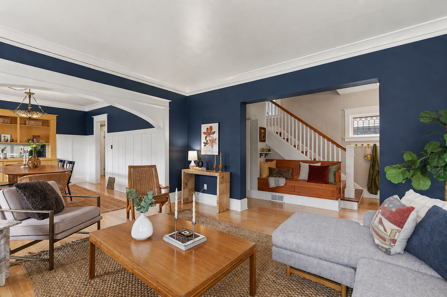 Interior view of a living room with blue walls, a wooden coffee table, a gray couch with colorful pillows, a beige armchair, and a wooden bench near a staircase, with natural light and decorative plants.