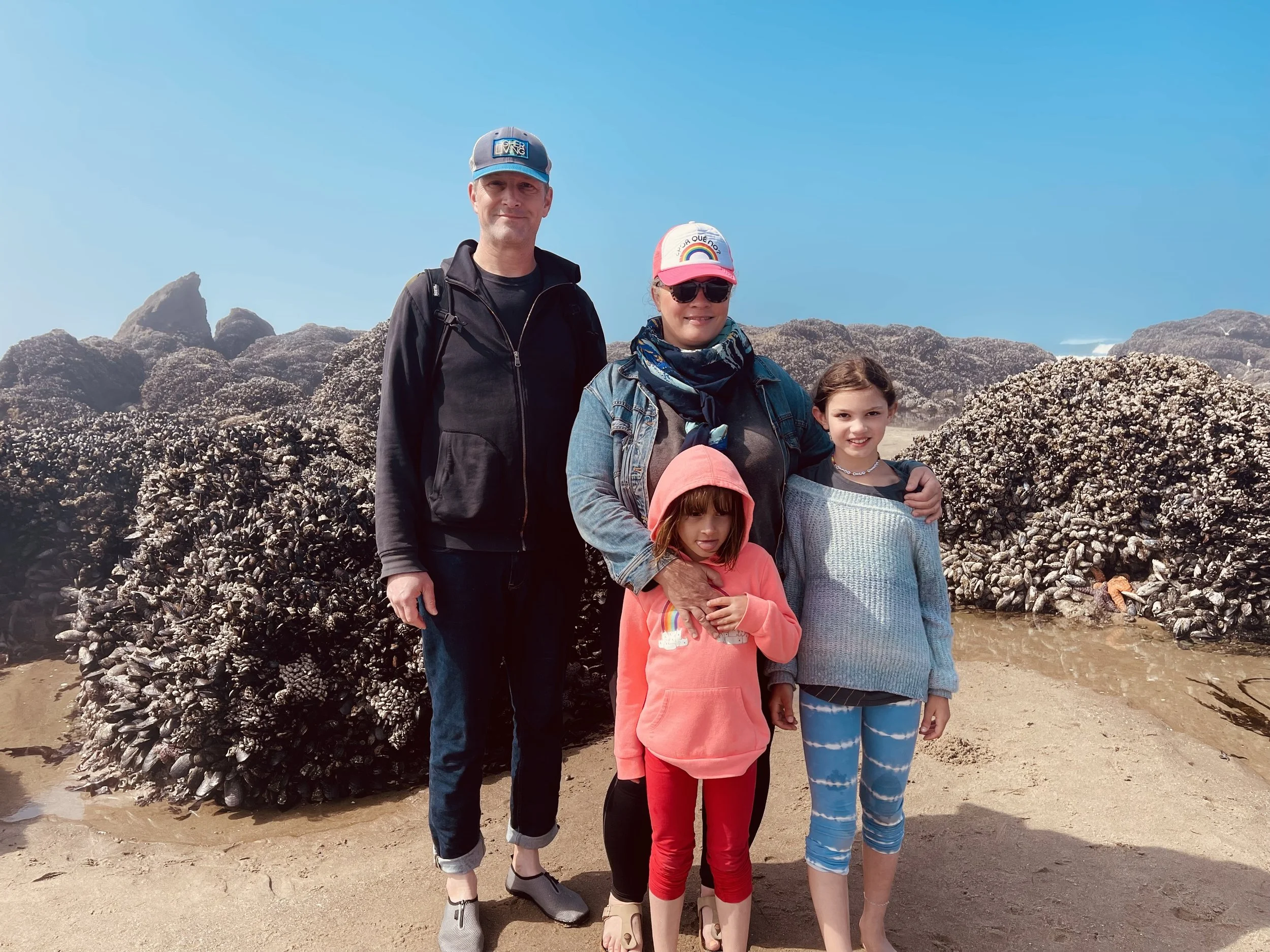 Family of five standing on the beach in front of a large cluster of mussel shells, with rocky formations and blue sky in the background.