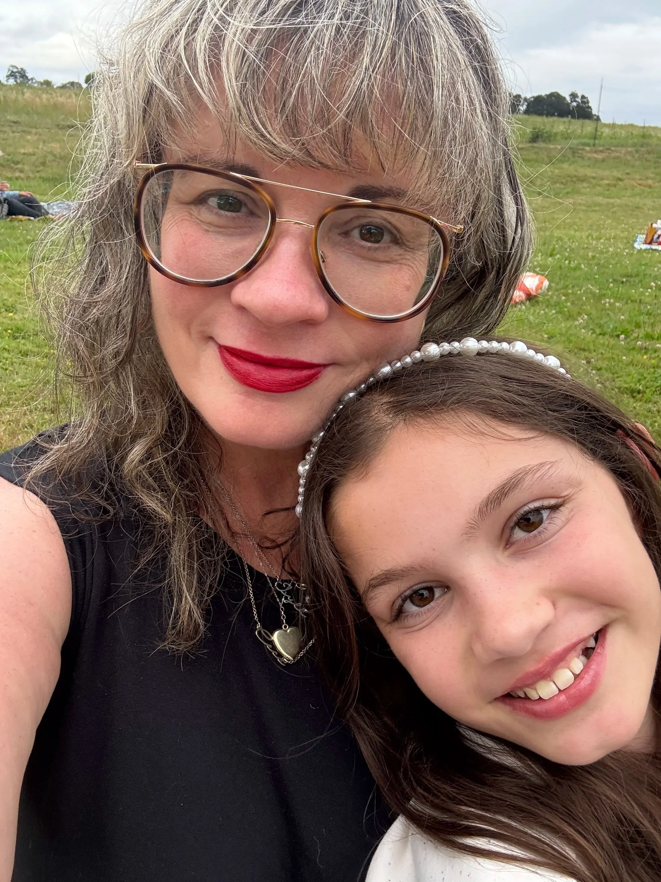Close-up of a woman with gray hair, glasses, makeup, and red lipstick, smiling outdoors with a young girl with brown hair, pearl headband, and brown eyes, in a grassy field.