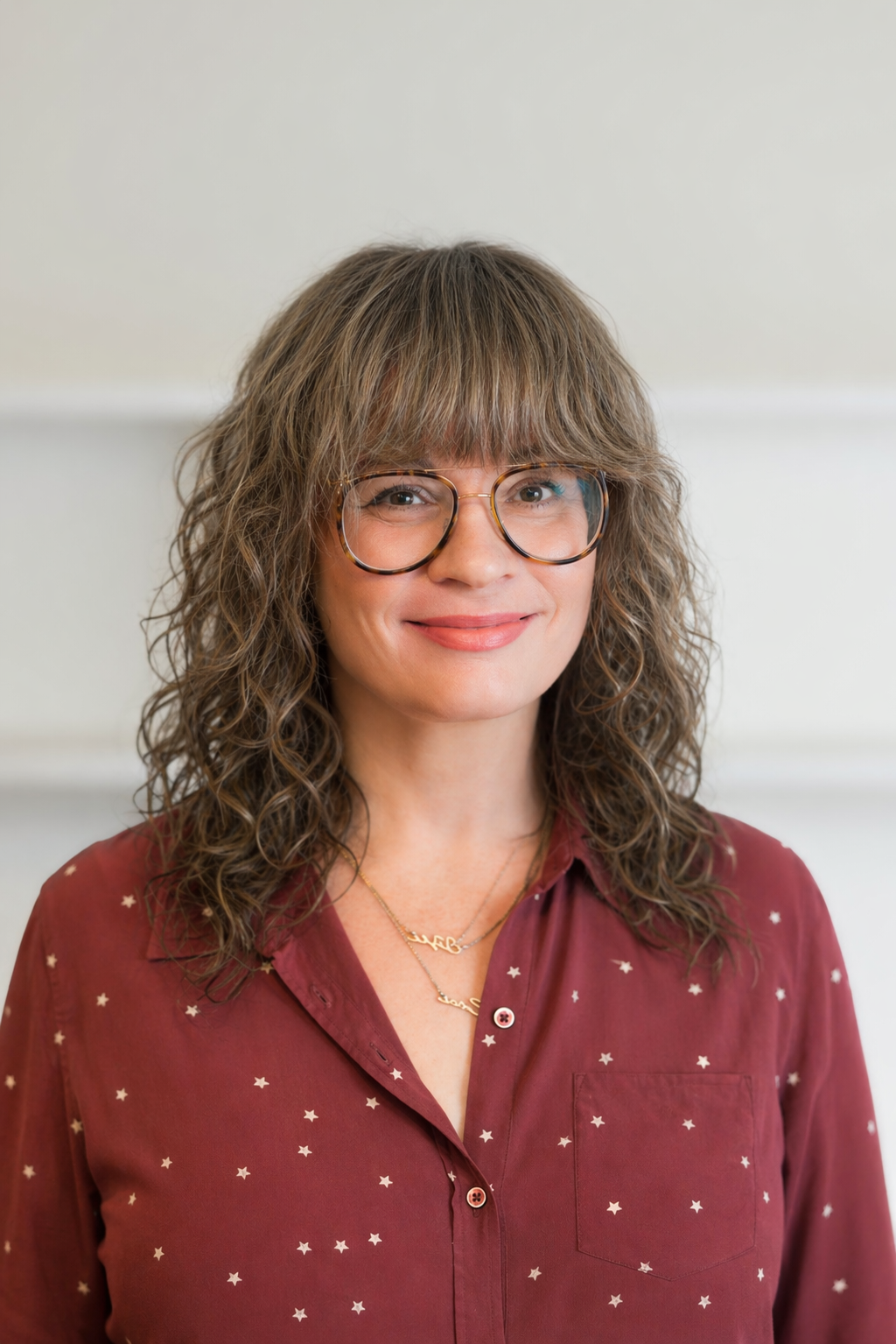 A woman with curly brown hair and glasses, wearing a maroon shirt with small white star patterns, standing indoors.
