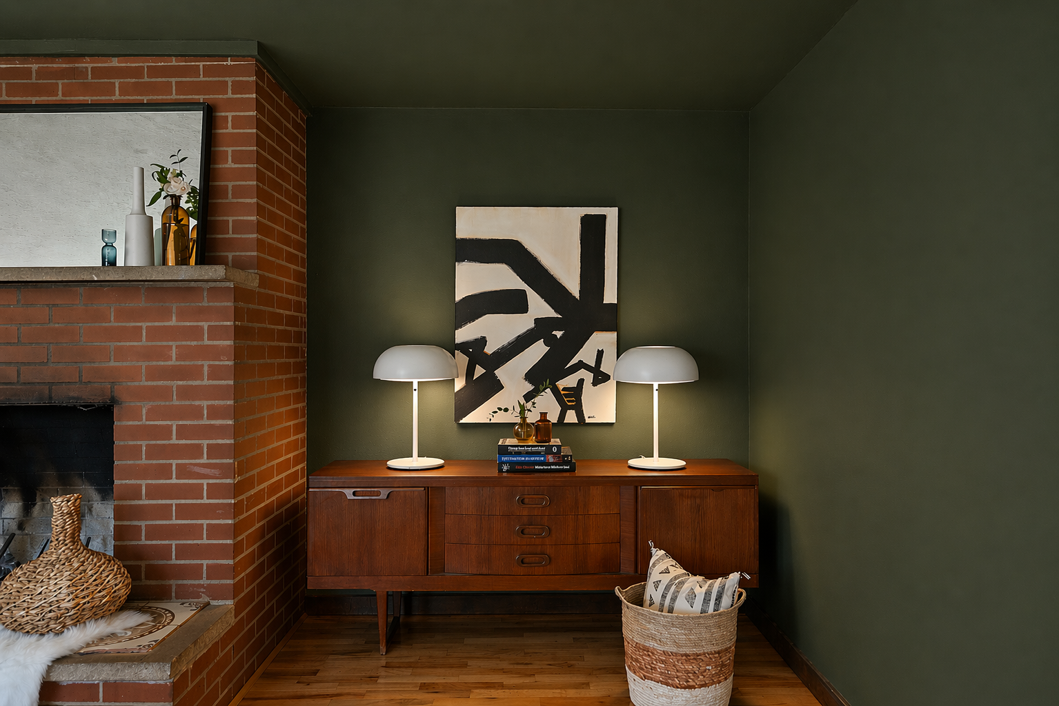 A mid-century modern wooden sideboard with two white lamps, a stack of books, and small decorative items against a dark green wall. An abstract black and white painting hangs above the sideboard. To the right, a woven basket with pillows, and to the left, a fireplace with a brick chimney and a woven basket with a fluffy white blanket.
