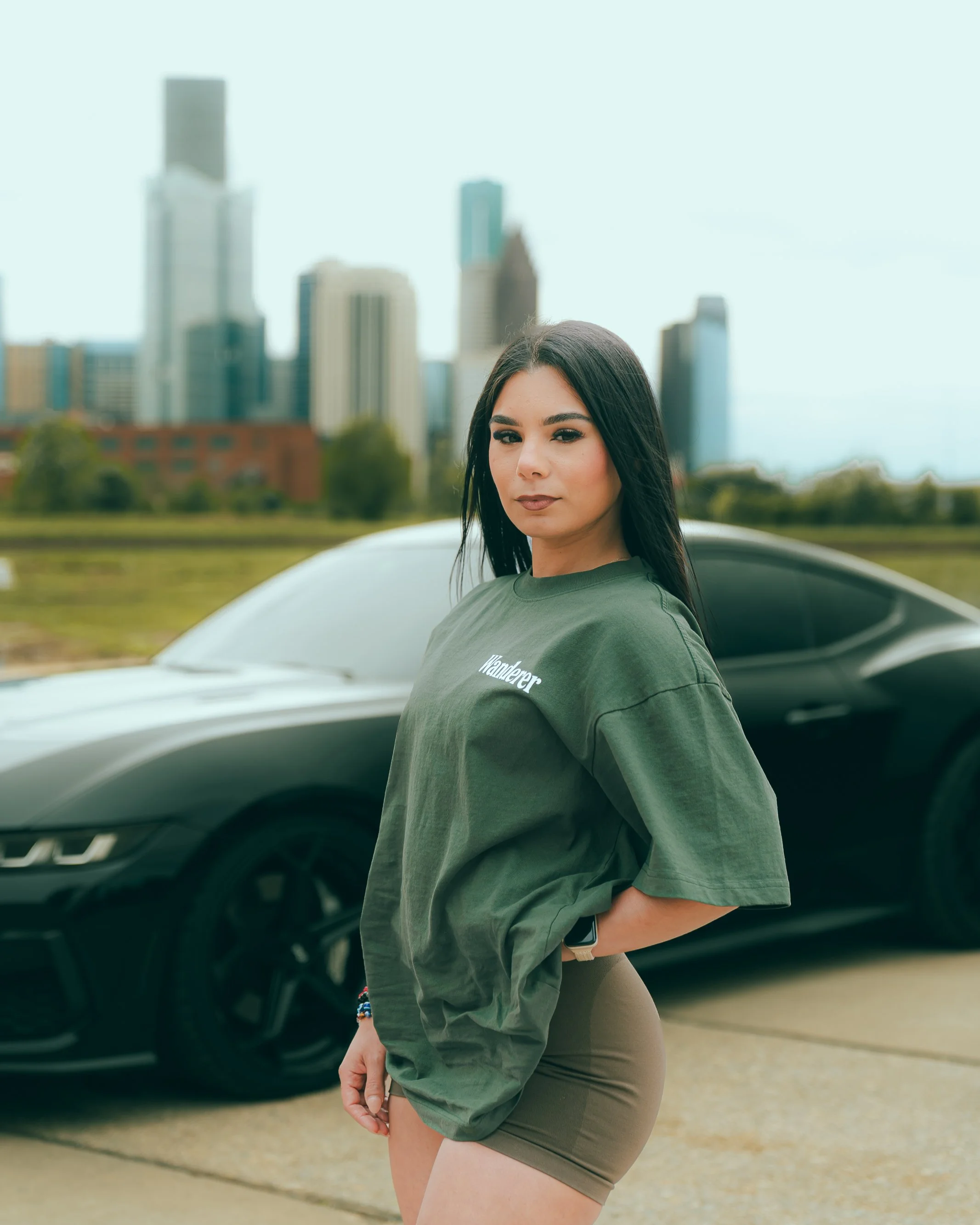 A woman with long dark hair and olive skin, wearing a green T-shirt and beige shorts, standing outdoors with a cityscape and tall buildings in the background, leaning against a white car.