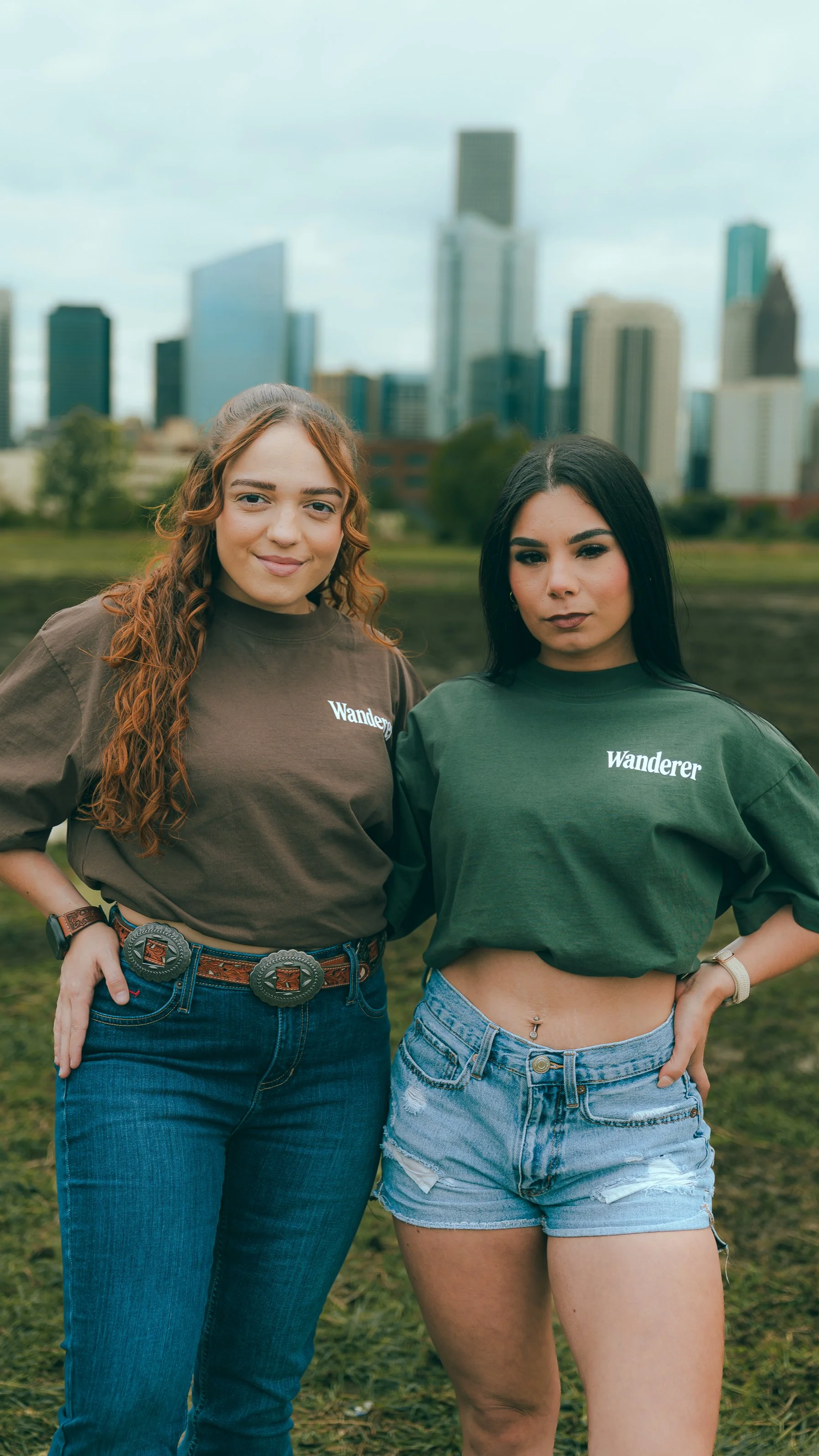 Two women standing outdoors in front of a city skyline, one with curly red hair and the other with straight black hair, both wearing green t-shirts with 'Wanderer' written on them.