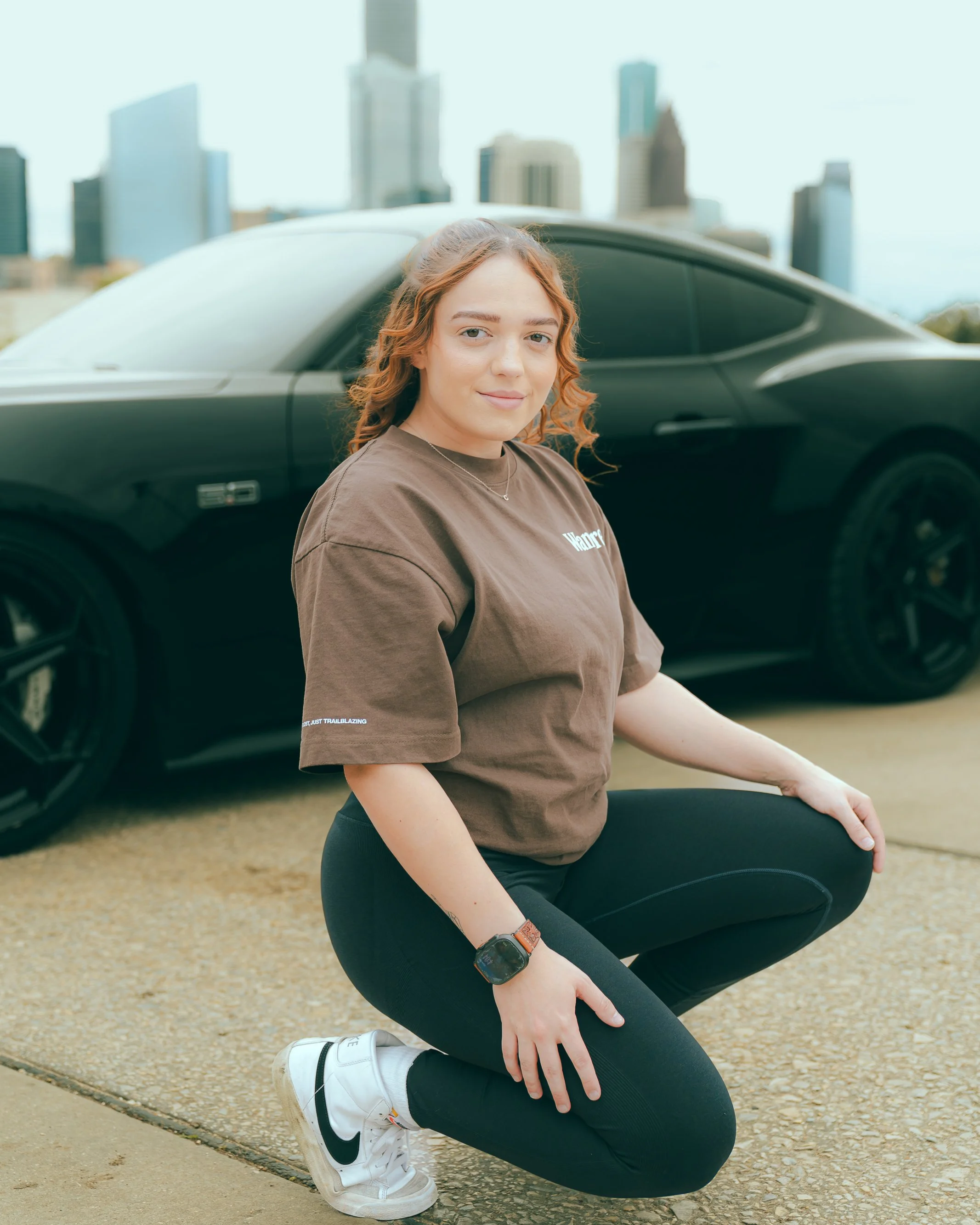 A woman with red hair and a small smile, wearing a brown t-shirt, black leggings, and white Nike sneakers, crouching on a concrete sidewalk in front of a black car with a city skyline in the background.